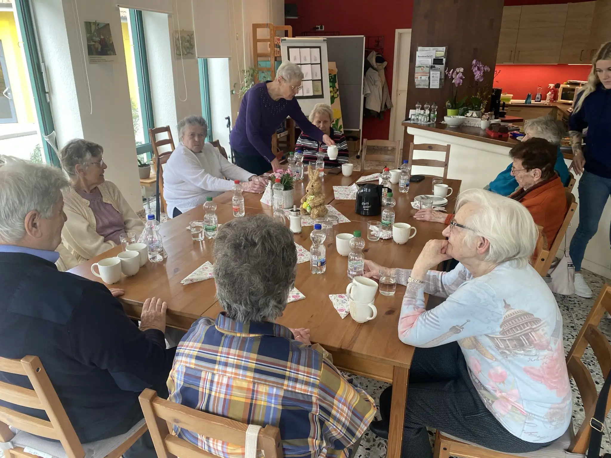 Eine Gruppe von Senioren sitzt an einem großen Holztisch und genießt gemeinsam Kaffee und Getränke in einem hellen Gemeinschaftsraum. Eine Betreuerin oder Angehörige steht am Tisch und schenkt etwas ein.