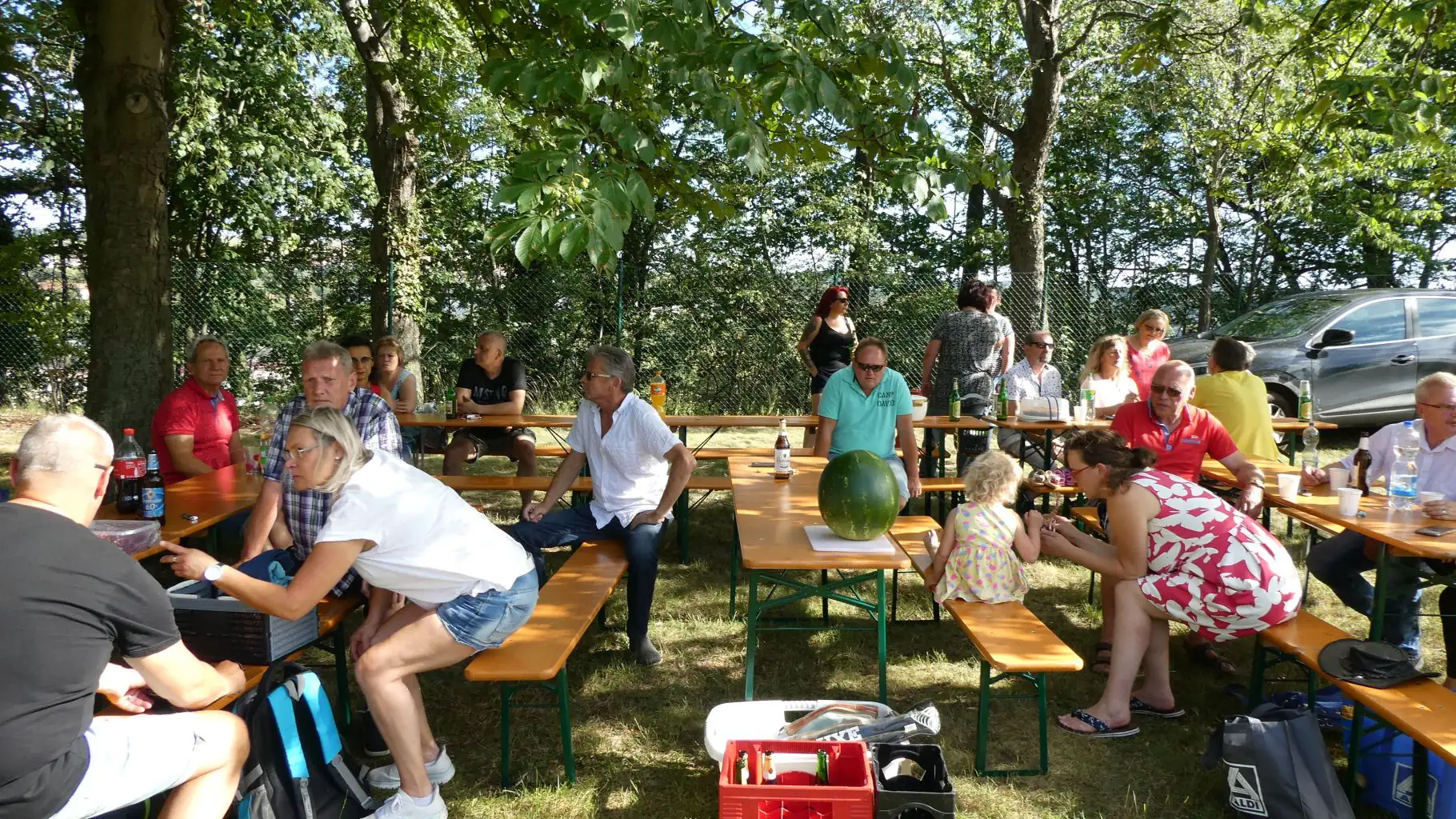 Eine Gruppe von Menschen sitzt bei sonnigem Wetter an Tischen im Freien und verbringt Zeit zusammen.