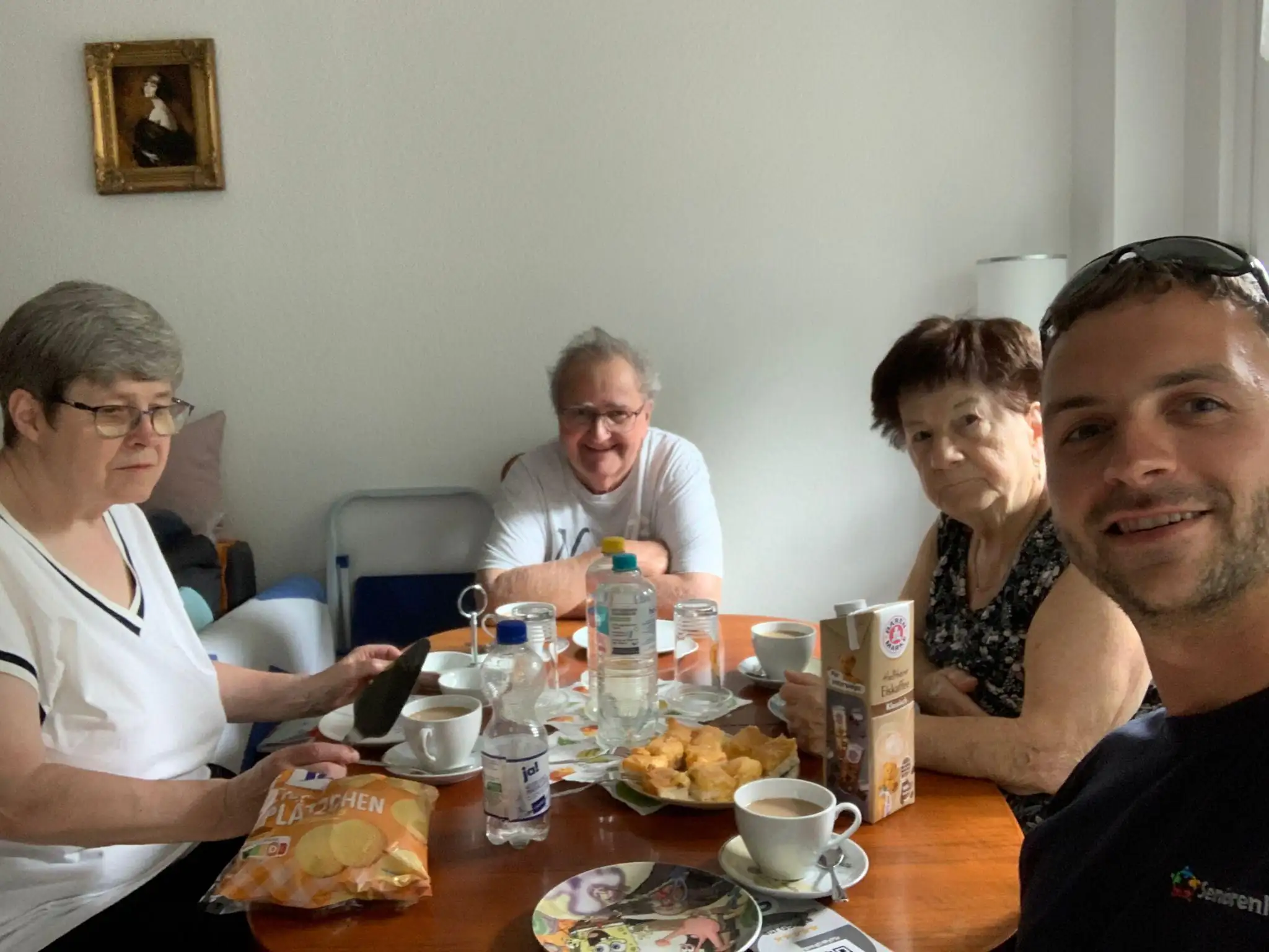 Eine Gruppe von Senioren und ein Betreuer sitzen gemeinsam am Esstisch und genießen Kaffee und Snacks.