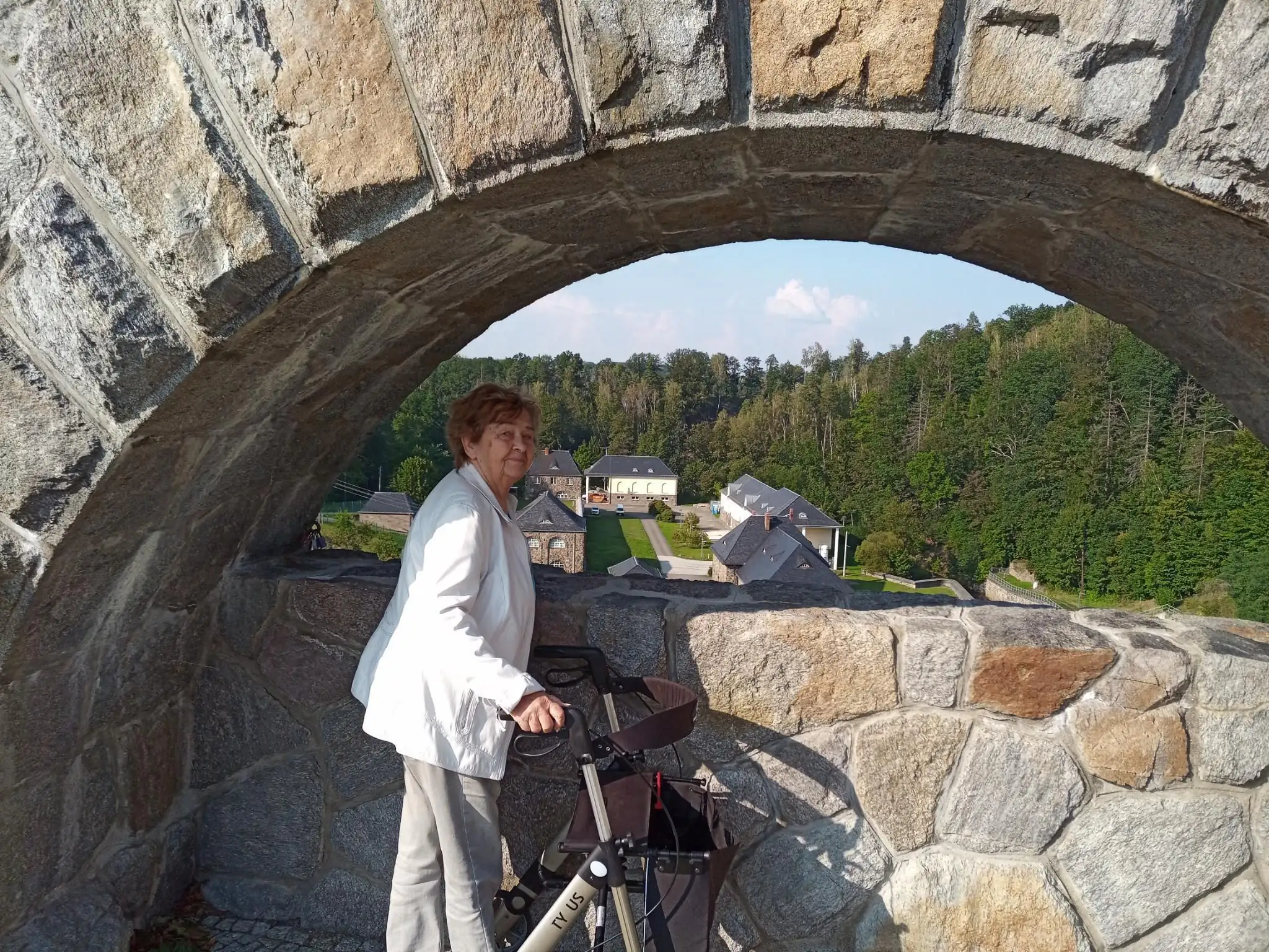 Eine ältere Dame steht mit einem Rollator unter einem steinernen Bogen und blickt auf eine ländliche Siedlung im Hintergrund. Das Bild zeigt Aktivität und Mobilität im Alter in einer natürlichen Umgebung.