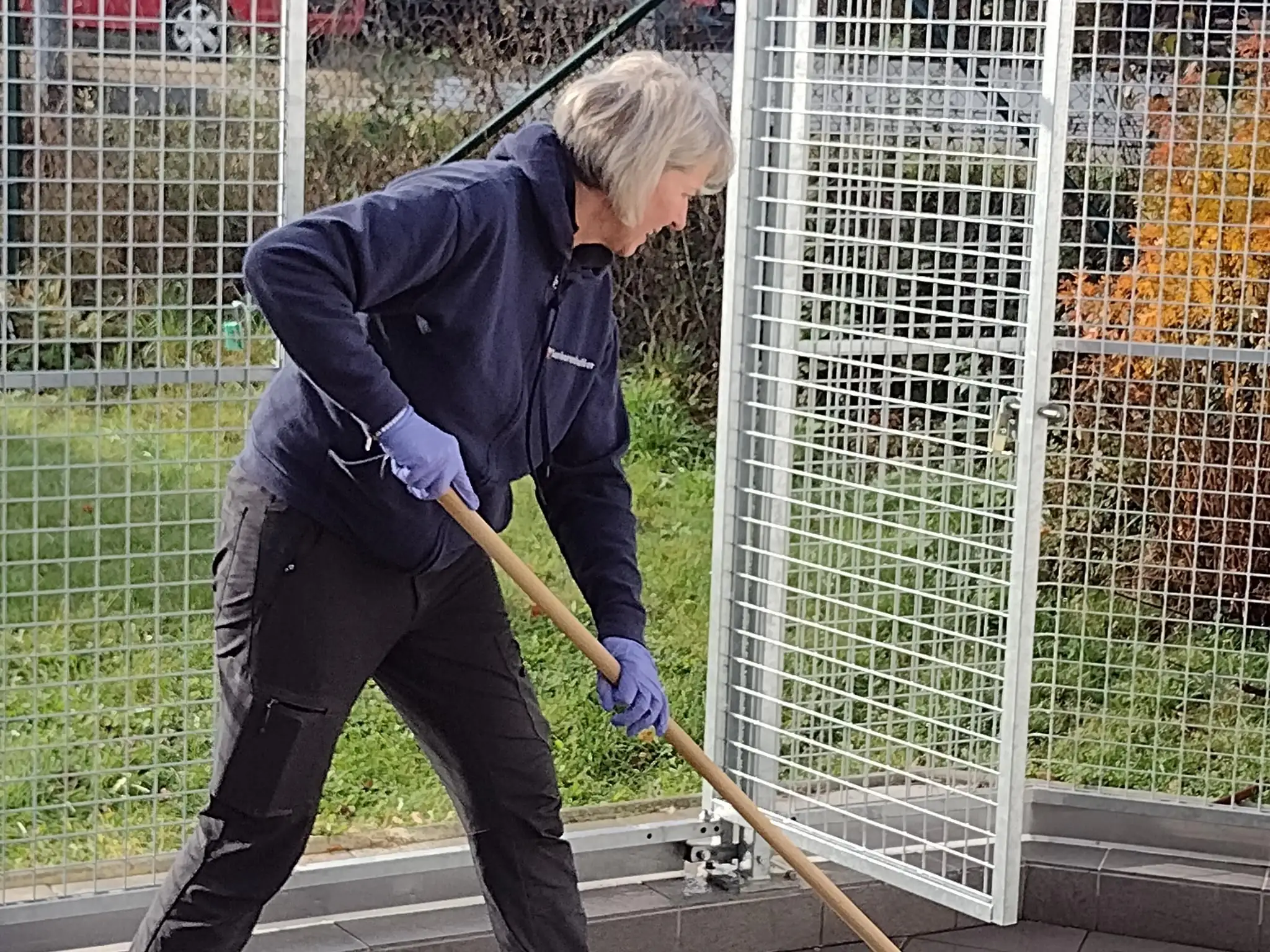 Eine Mitarbeiterin in Arbeitskleidung und Schutzhandschuhen kehrt den Außenbereich eines Geländes. Die Szene zeigt pflegerische oder hauswirtschaftliche Tätigkeiten im Freien.