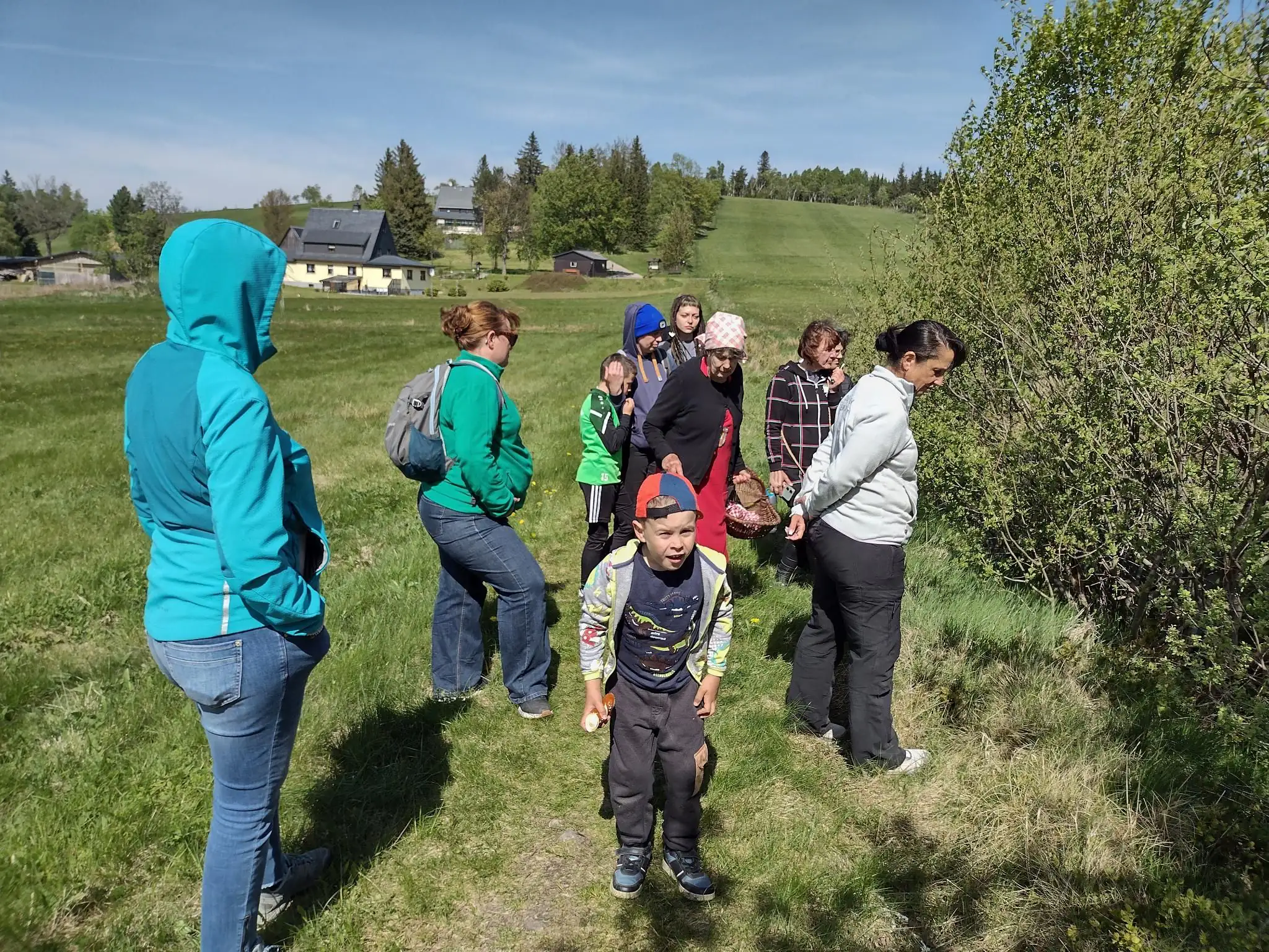 Eine gemischte Gruppe aus Erwachsenen und Kindern unternimmt einen Spaziergang auf einer grünen Wiese im Freien. Die Szene zeigt eine Freizeitaktivität in der Natur bei sonnigem Wetter.