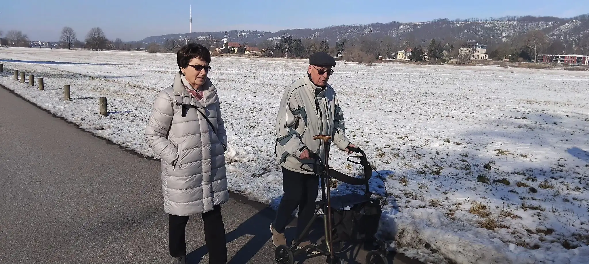 Zwei Senioren gehen bei winterlichen Bedingungen spazieren, wobei einer einen Rollator verwendet. Das Bild symbolisiert aktives Altern und Mobilitaet im Alter.