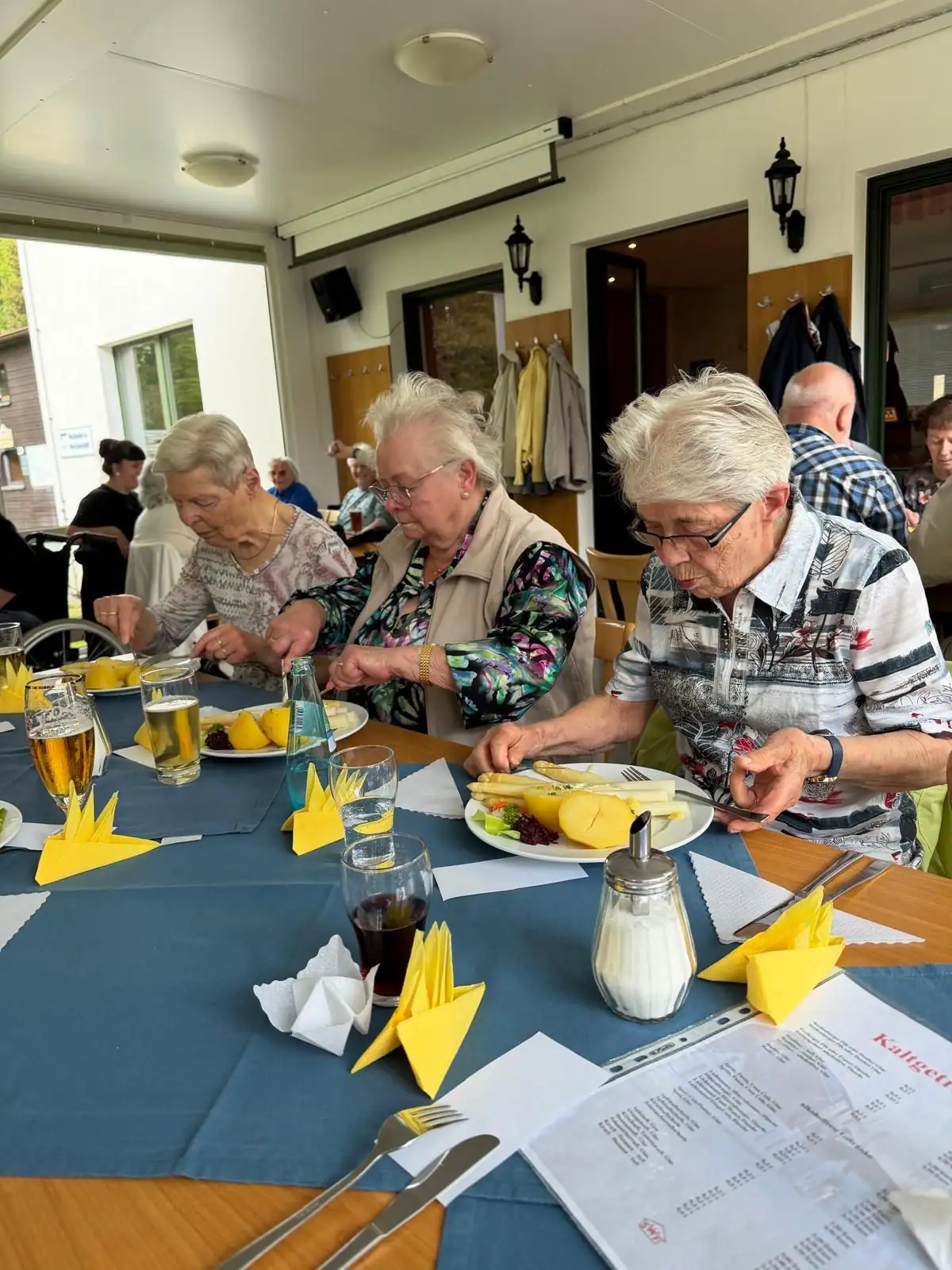 Drei ältere Damen sitzen an einem gedeckten Tisch und essen gemeinsam eine Mahlzeit. Im Vordergrund sind Getränke, Besteck und gefaltete Servietten zu sehen.