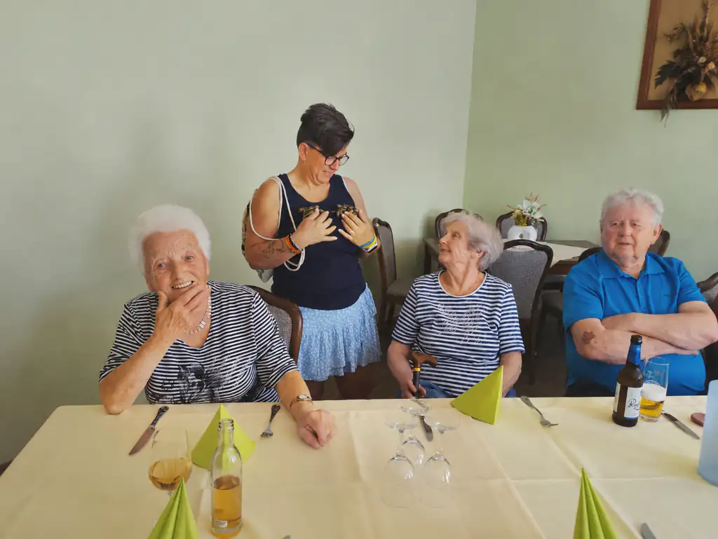 Eine Gruppe von Senioren und eine Betreuerin sitzen an einem gedeckten Tisch, wobei die Betreuerin zwei kleine Vögel hält. Die Szene zeigt eine gesellige Runde mit Tieren im Pflegekontext.