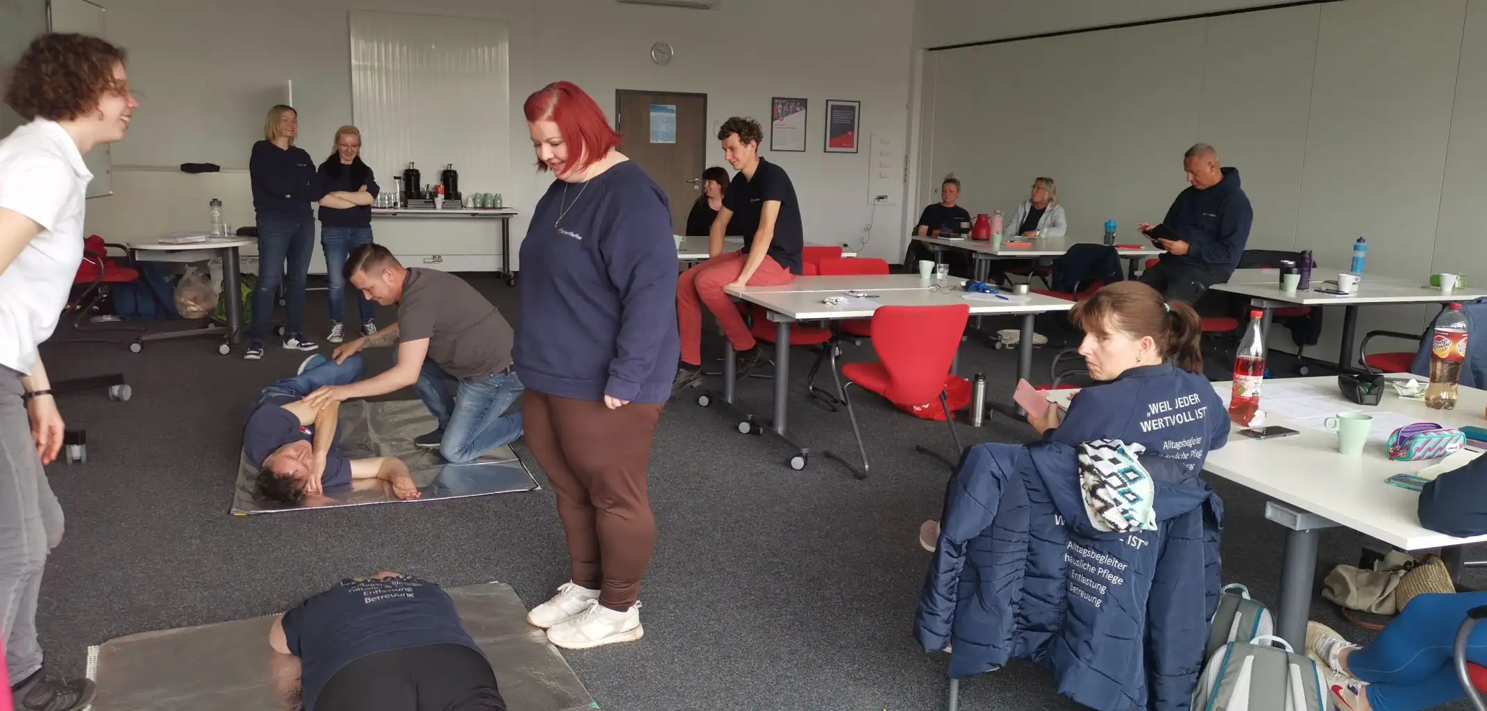 Schulungssituation in einem Seminarraum, bei der Pflegekräfte praktische Übungen auf Matten durchführen. Mehrere Personen beobachten die Demonstration von Techniken am Boden.