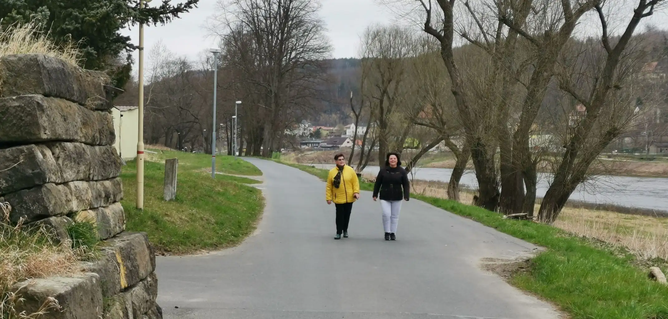 Zwei Frauen gehen gemeinsam auf einem asphaltierten Weg an einem Flussufer spazieren. Die Szene zeigt eine aktive Bewegung im Freien bei bewölktem Himmel.