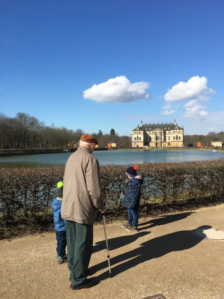 Ein älterer Herr mit Gehstock genießt einen sonnigen Tag im Park mit zwei Kindern vor einem historischen Schloss.
