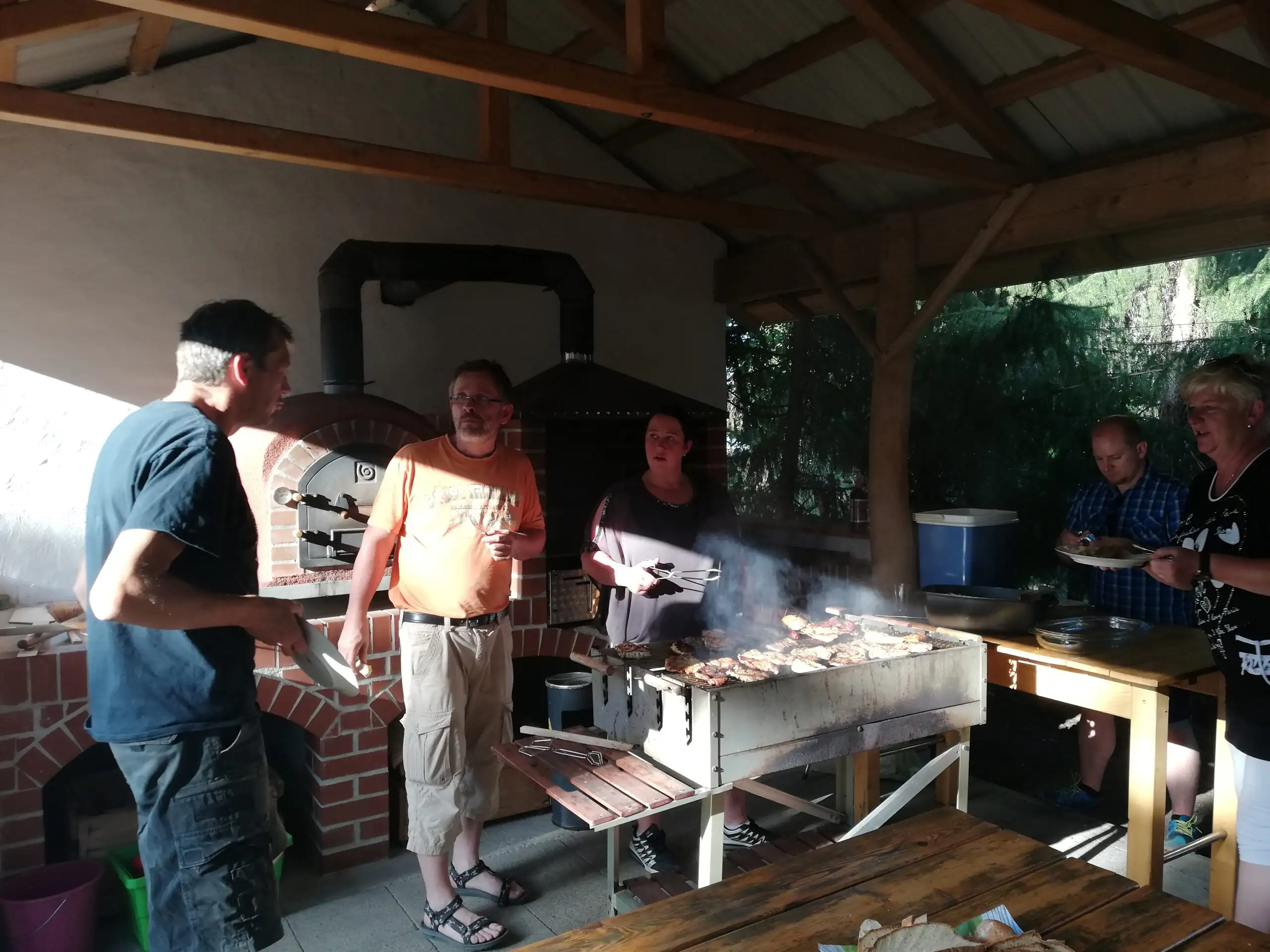 Eine Gruppe von Menschen steht bei einem Grillfest unter einer Holzüberdachung und bedient sich am Essen. Die Szene wirkt informell und zeigt eine gesellige Zusammenkunft im Freien.