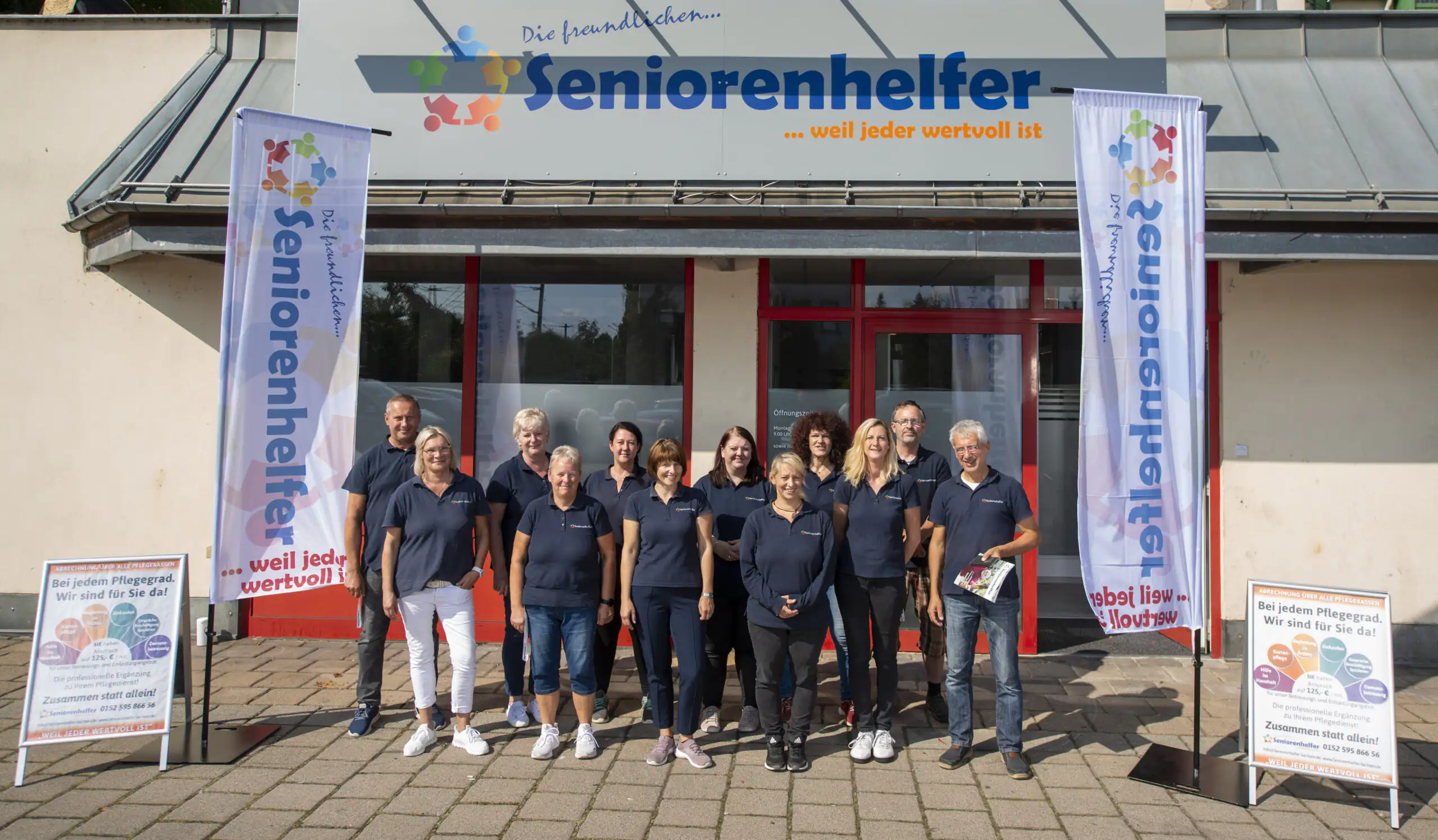 Gruppenfoto des Mitarbeiter-Teams von Seniorenhelfer vor dem Firmengebäude mit sichtbaren Markenbannern und Schildern.