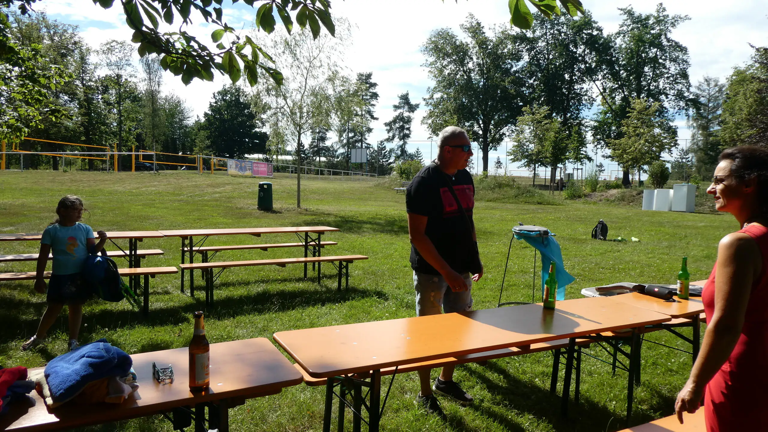Eine Gruppe von Menschen trifft sich bei sonnigem Wetter im Park an Picknicktischen. Im Hintergrund sind Bäume, Gras und ein Volleyballnetz zu sehen.