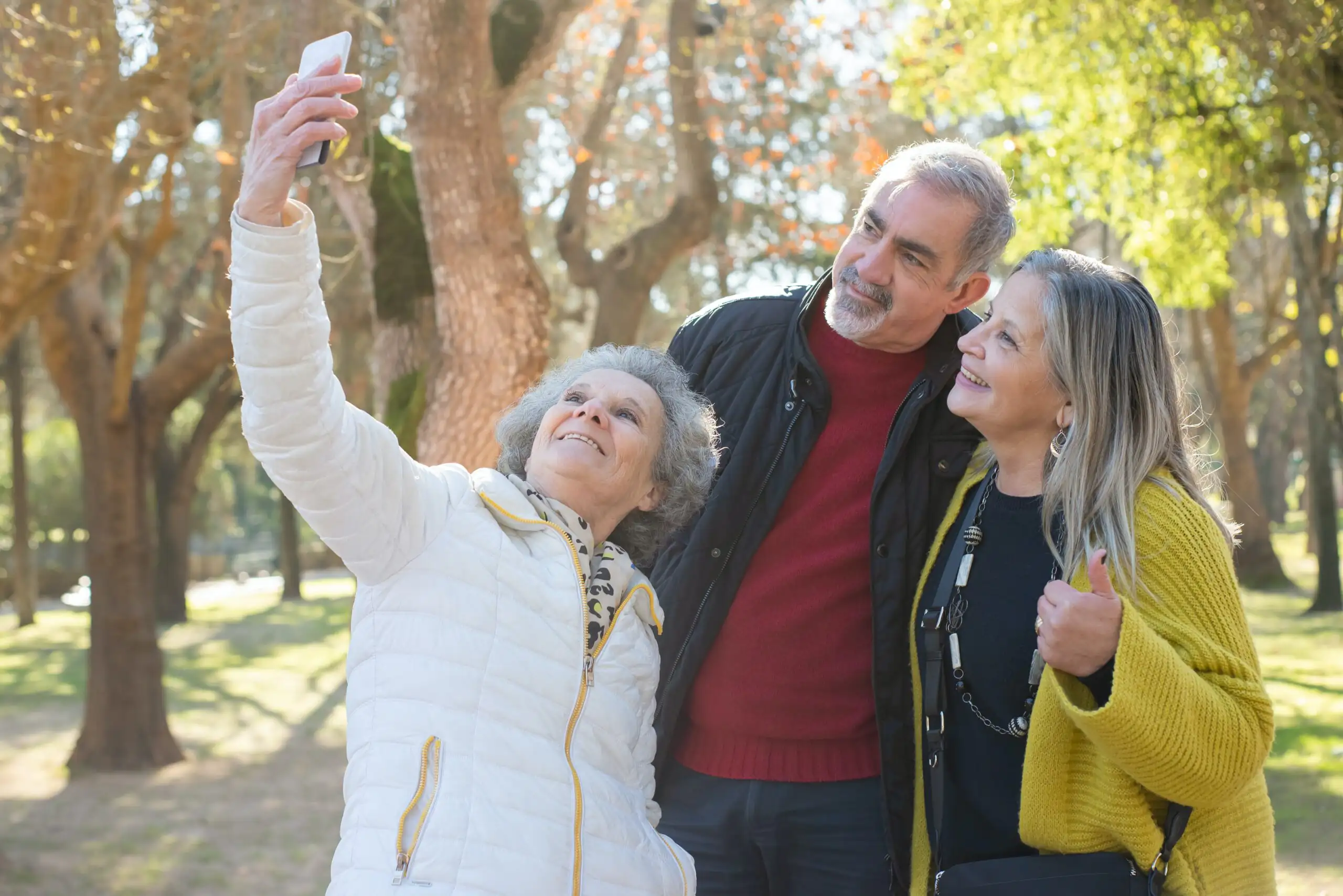 Drei ältere Menschen machen gemeinsam ein Selfie im Park bei sonnigem Wetter. Die Gruppe wirkt glücklich und aktiv im Freien.