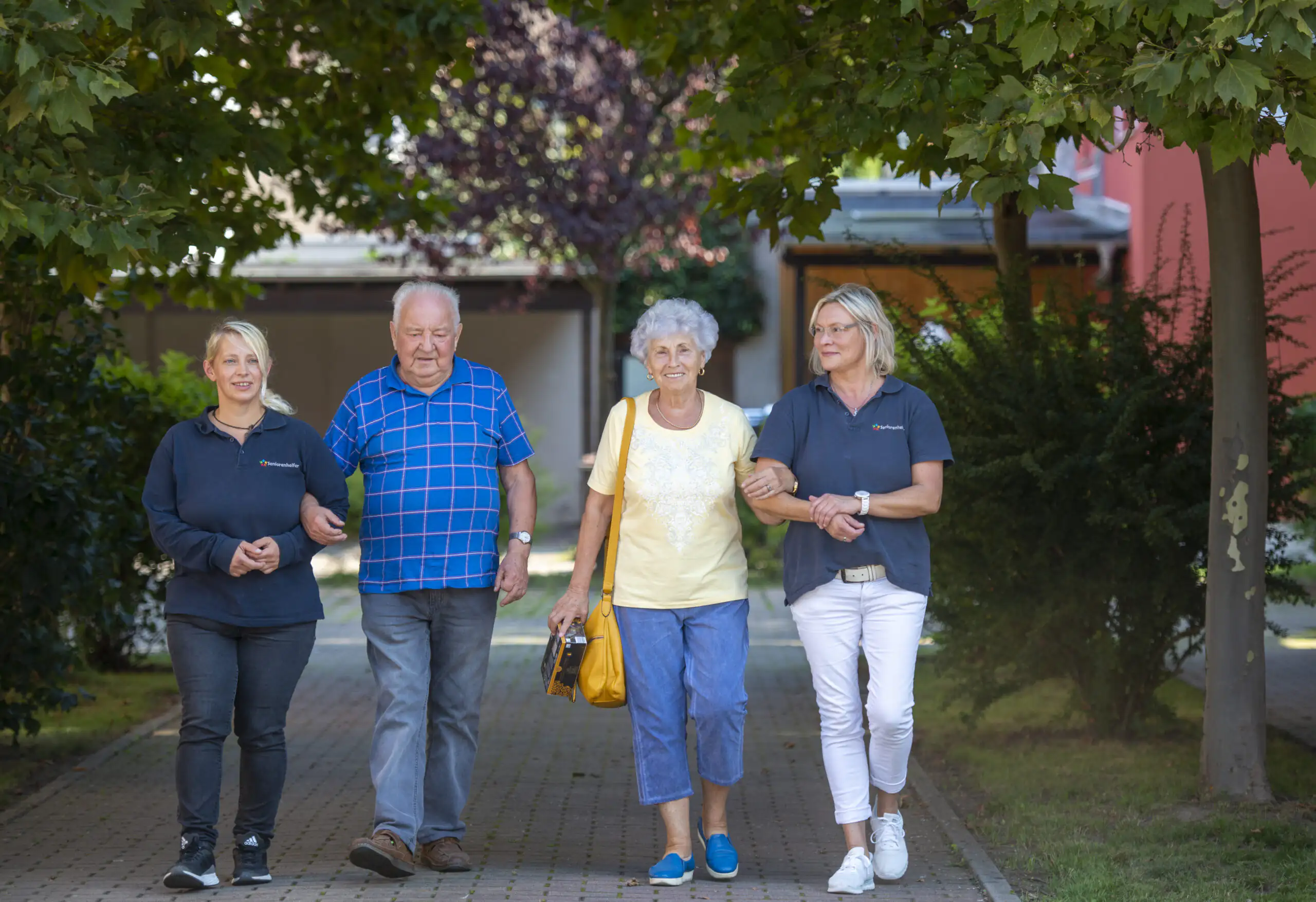 Zwei Pflegekräfte in Uniform begleiten zwei Senioren bei einem Spaziergang im Freien. Die Gruppe geht arm in arm auf einem gepflasterten Weg unter Bäumen.