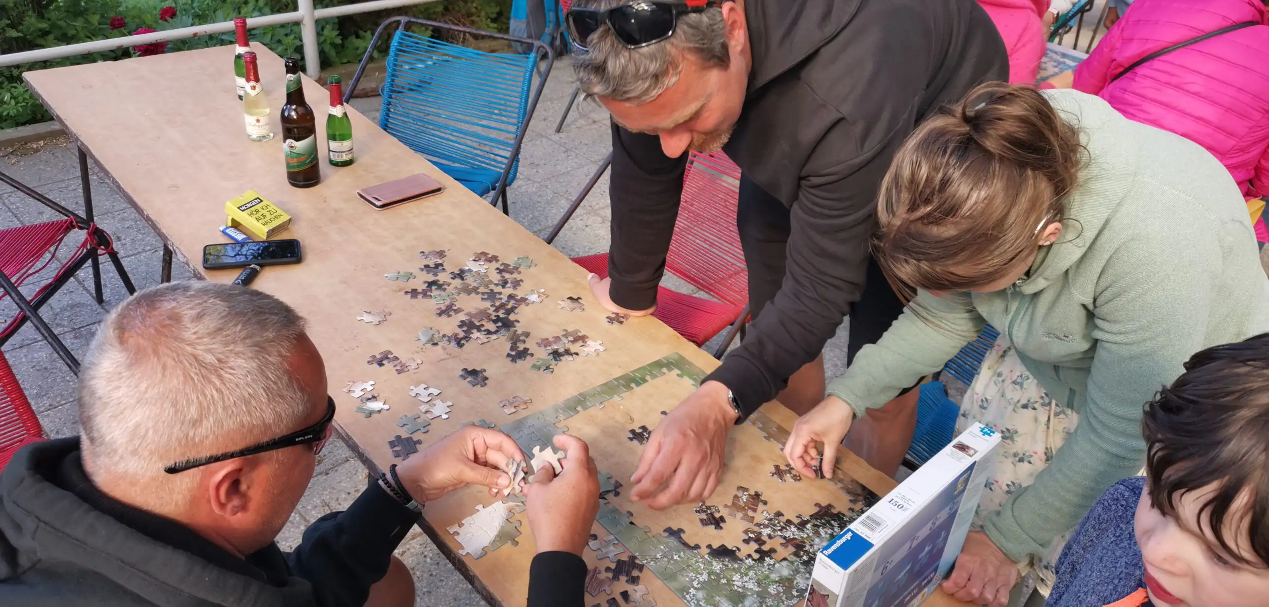 Eine Gruppe von Menschen arbeitet gemeinsam an einem Puzzlespiel an einem Tisch im Freien. Im Hintergrund sind Getränkeflaschen und persönliche Gegenstände auf dem Tisch sichtbar.
