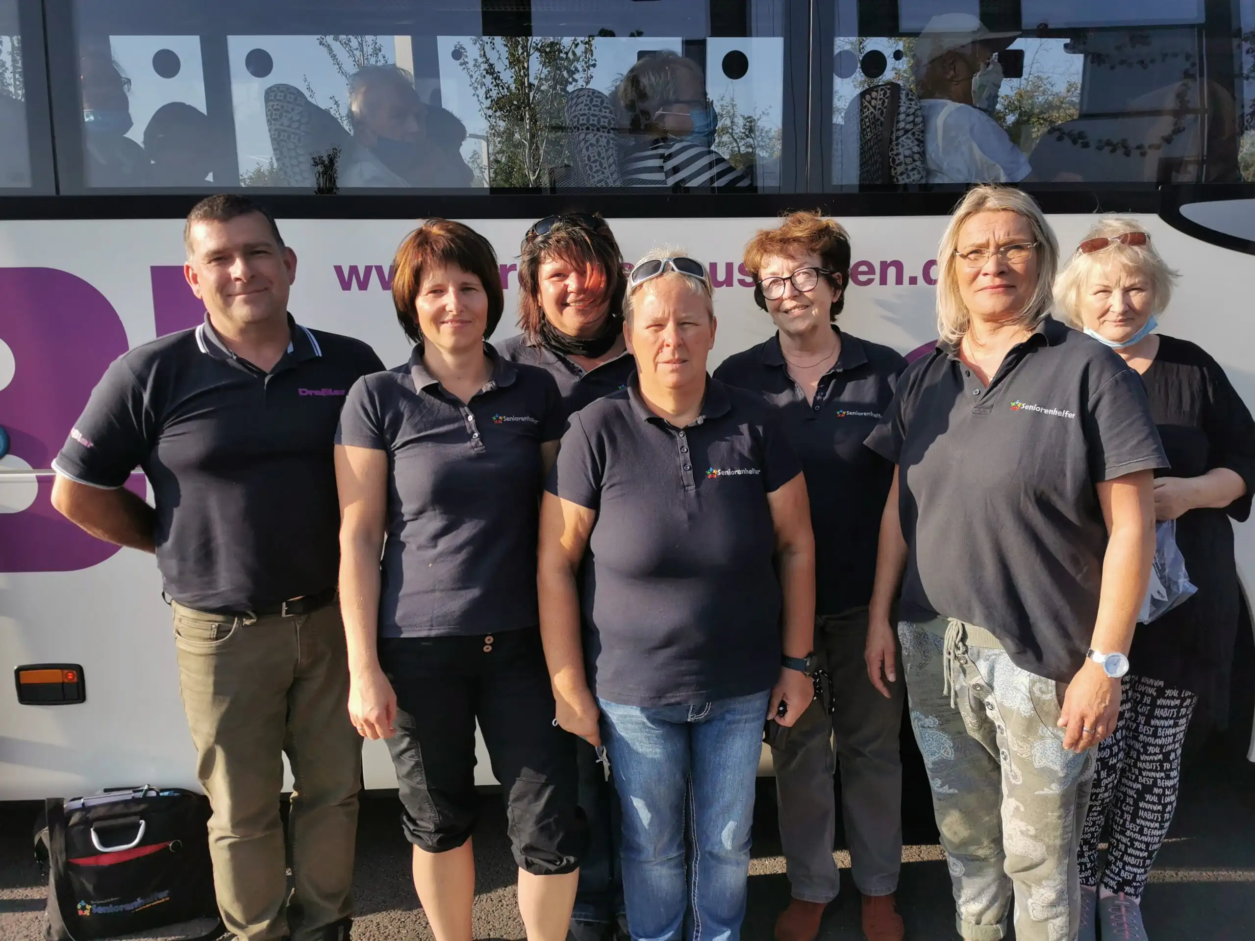 Eine Gruppe von sieben Personen in einheitlicher Arbeitskleidung steht vor einem weißen Bus. Im Fenster des Busses sind undeutliche Spiegelungen weiterer Personen zu erkennen.