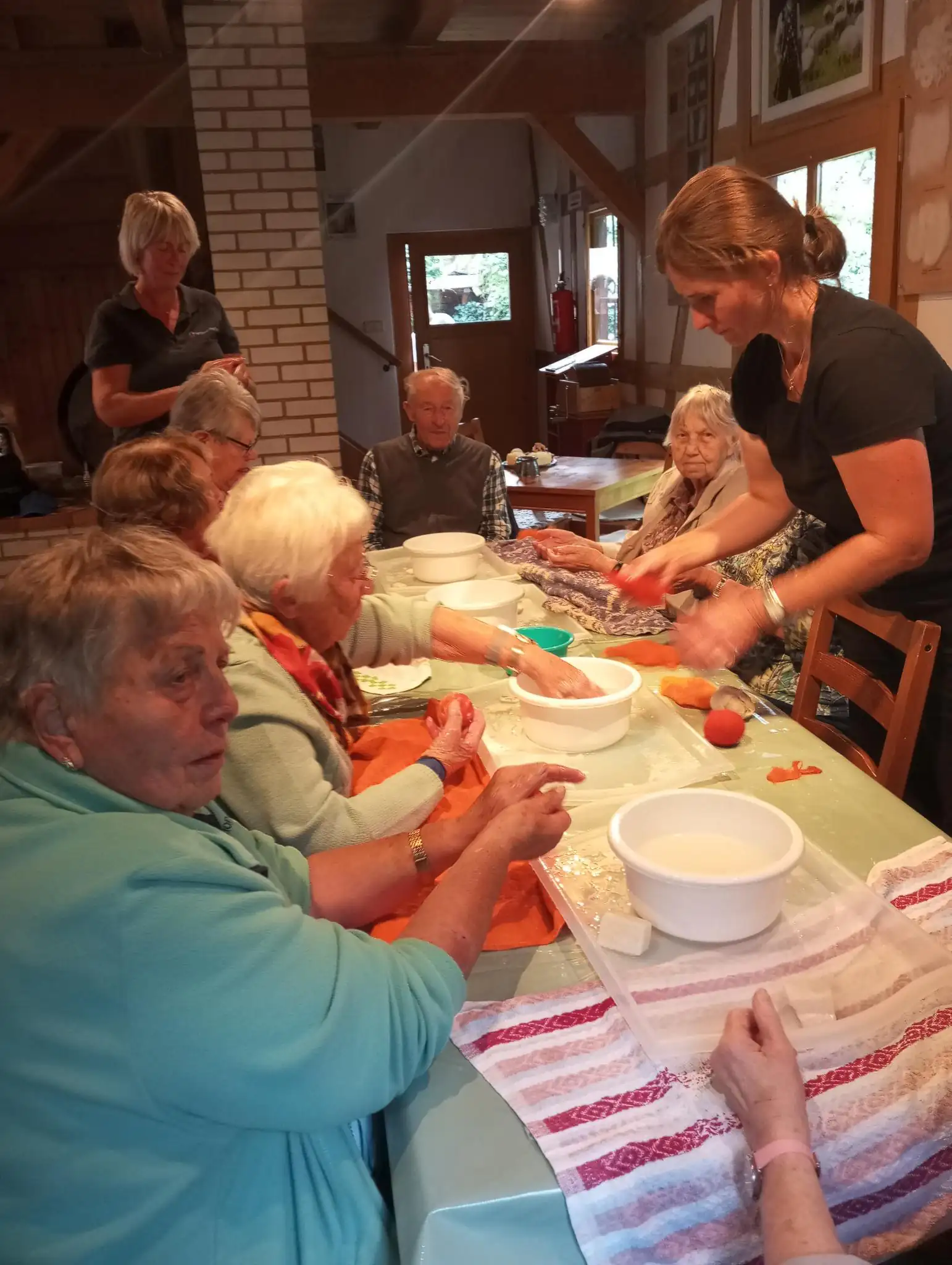 Eine Gruppe von Senioren nimmt an einer kreativen Aktivität am Tisch teil. Eine Betreuungskraft unterstützt die Teilnehmer bei der Beschäftigung.