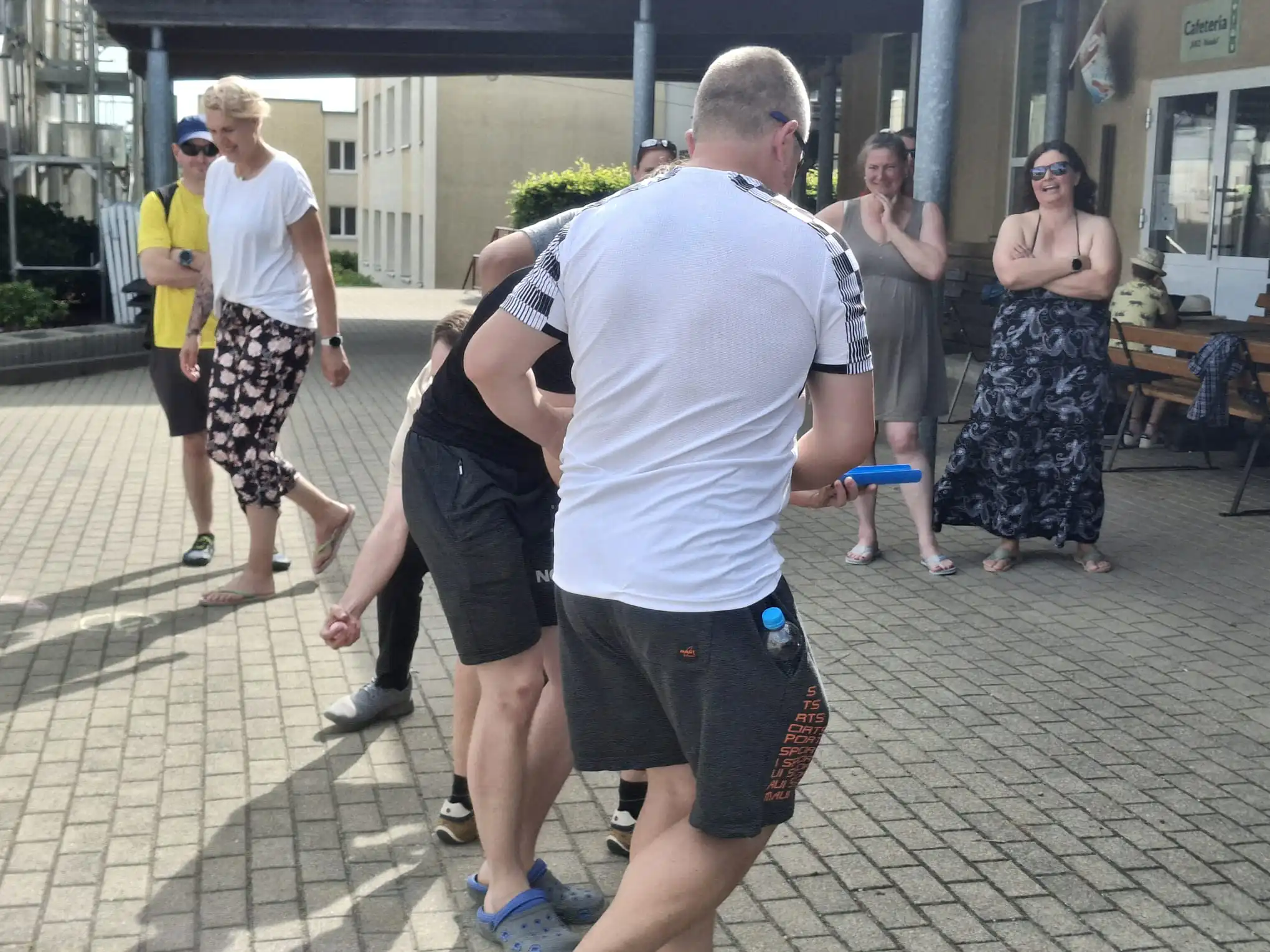 Eine Gruppe von Menschen spielt ein Spiel im Freien auf einem gepflasterten Hof. Die Szene wirkt wie eine informelle Freizeitaktivität oder ein Sommerfest.