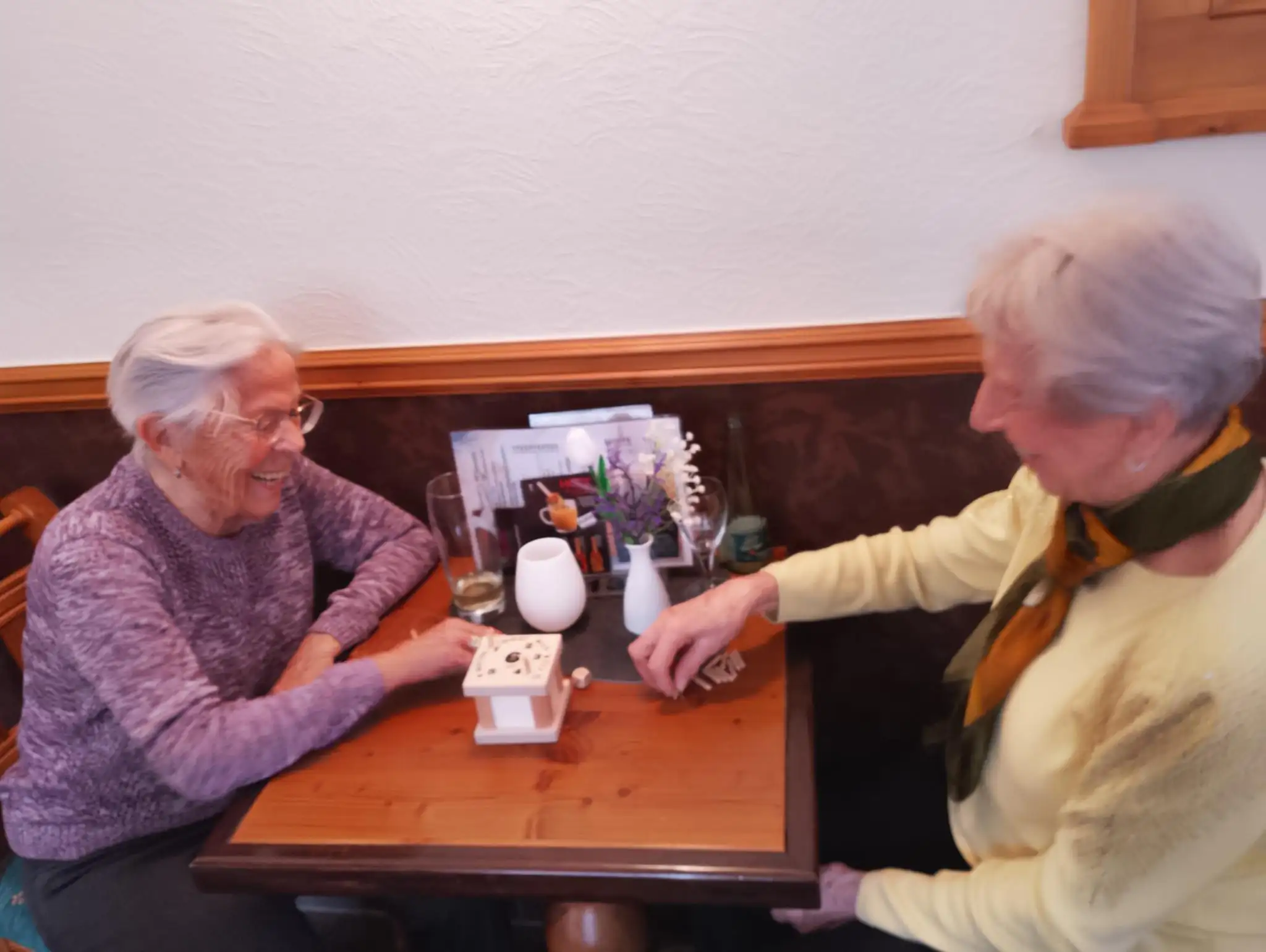 Zwei Seniorinnen spielen gemeinsam ein Brettspiel an einem Holztisch und unterhalten sich angeregt.