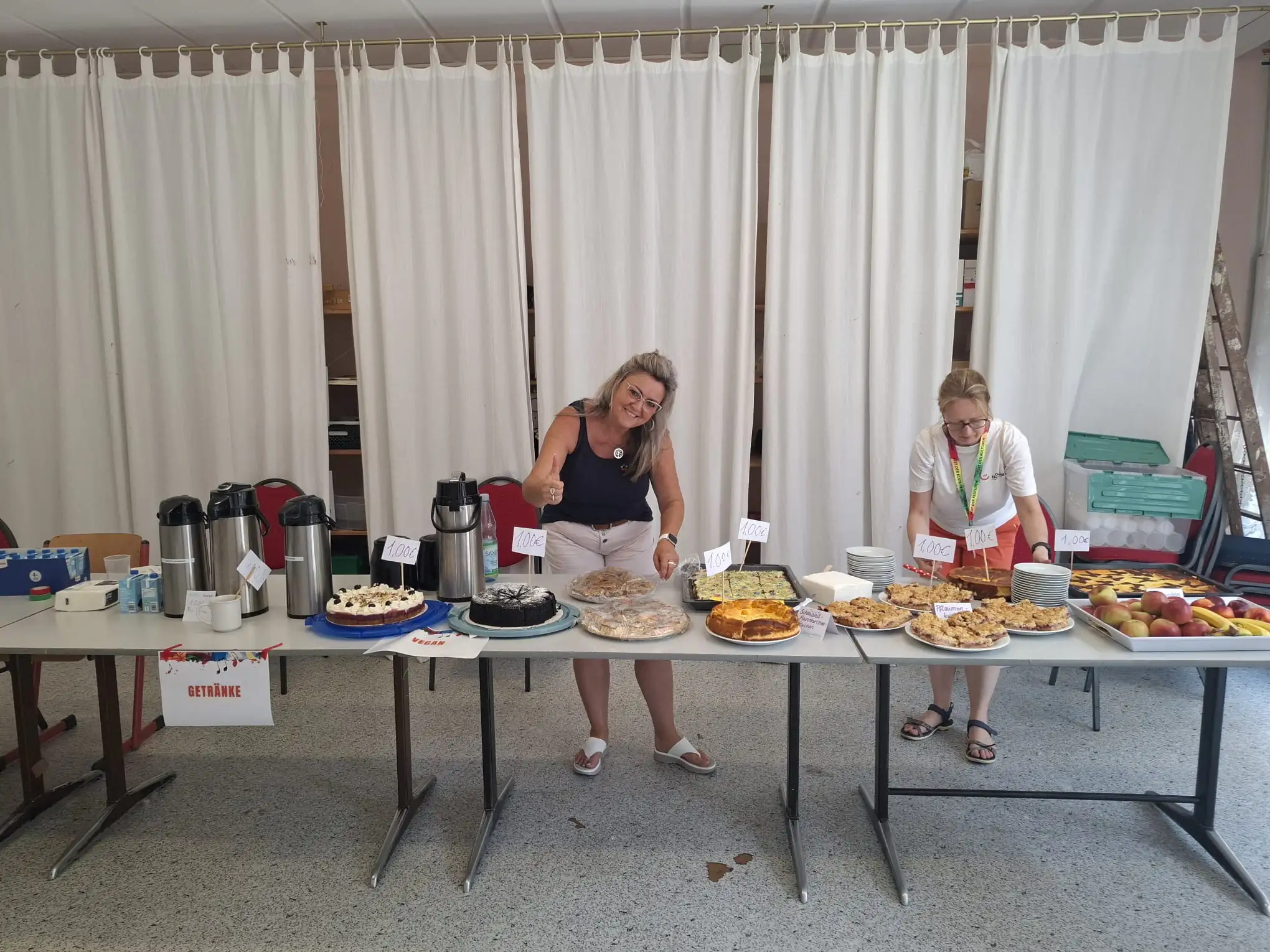 Zwei Frauen präsentieren ein reichhaltiges Angebot an selbstgebackenen Kuchen und Getränken bei einem Gemeinschaftsereignis. Die Szene strahlt eine freundliche und einladende Atmosphäre aus.