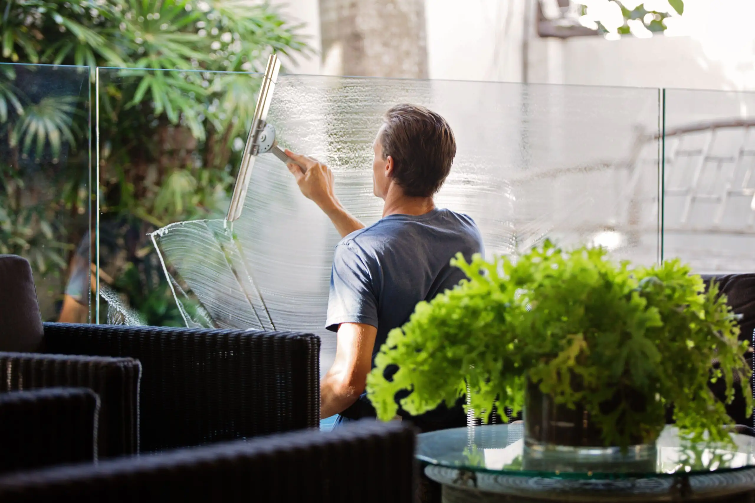 Ein Mann in blauem T-Shirt reinigt eine Glasbrüstung auf einer Terrasse mit Gartenmöbeln.