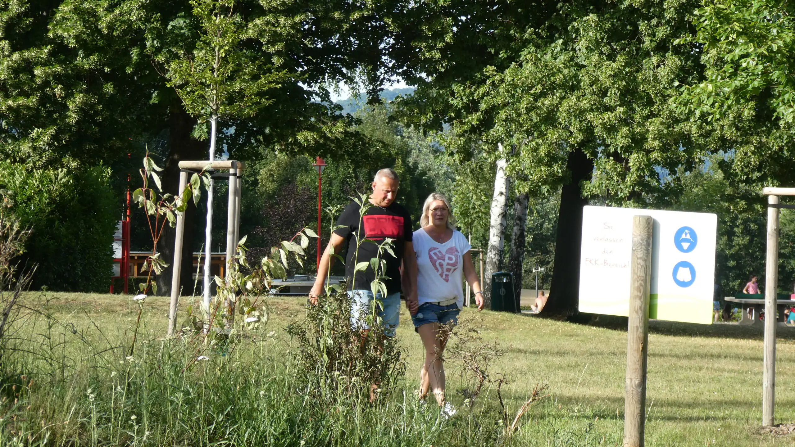 Ein Mann und eine Frau gehen Hand in Hand durch einen grünen Park im Sommer. Die Szene wirkt entspannt und zeigt Aktivität im Freien.