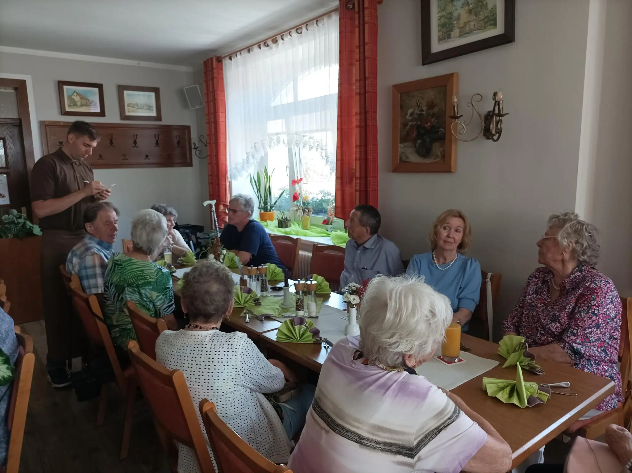 Eine Gruppe älterer Menschen sitzt gemeinsam an einem gedeckten Tisch und isst, während ein Betreuer Notizen macht.