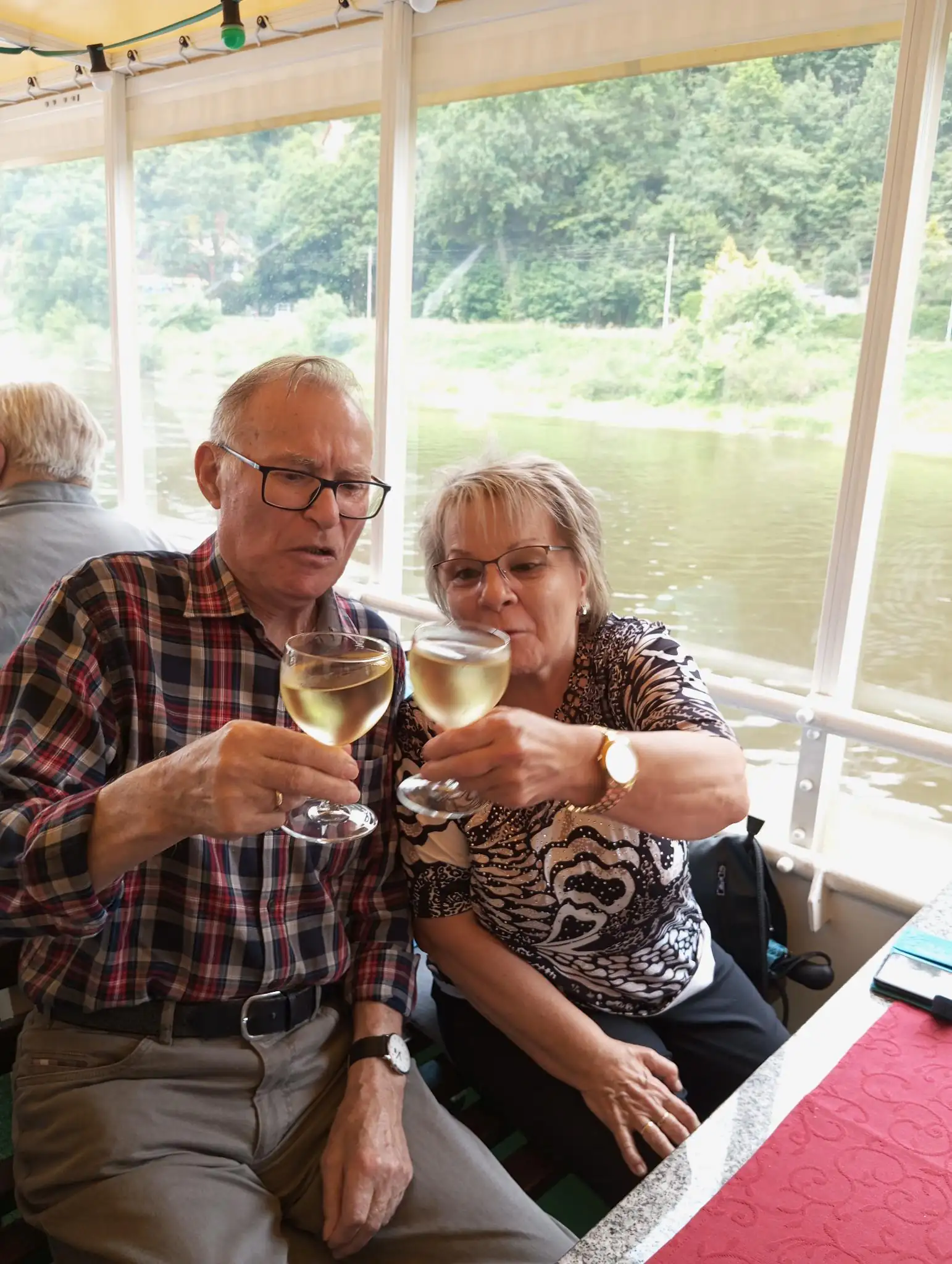 Ein älteres Paar genießt eine Flussfahrt und stößt dabei mit Gläsern Weißwein an.