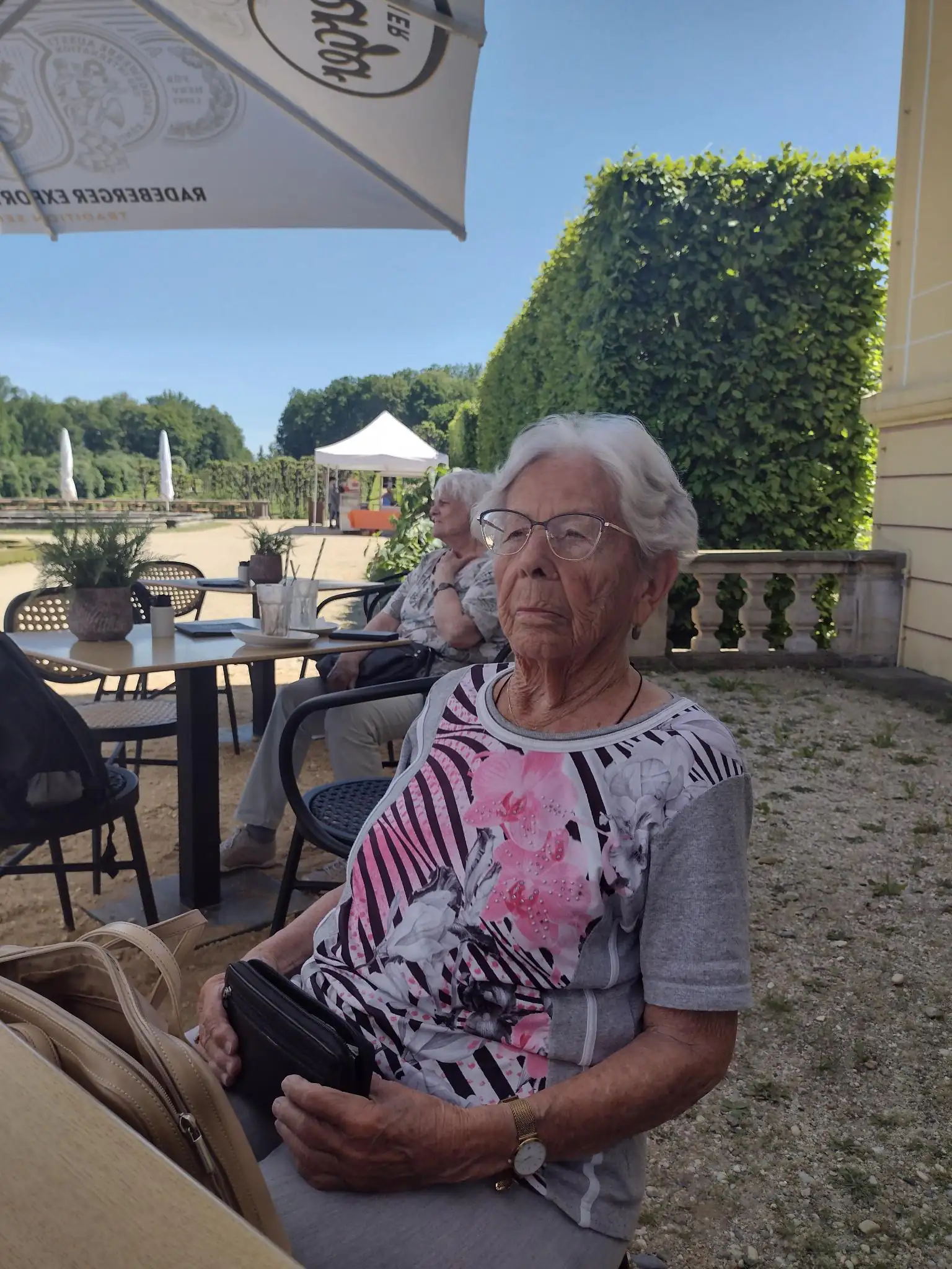 Eine ältere Dame mit Brille sitzt im Freien an einem sonnigen Tag auf einer Terrasse oder in einem Park. Im Hintergrund sind weitere Personen und eine grüne Hecke zu sehen.