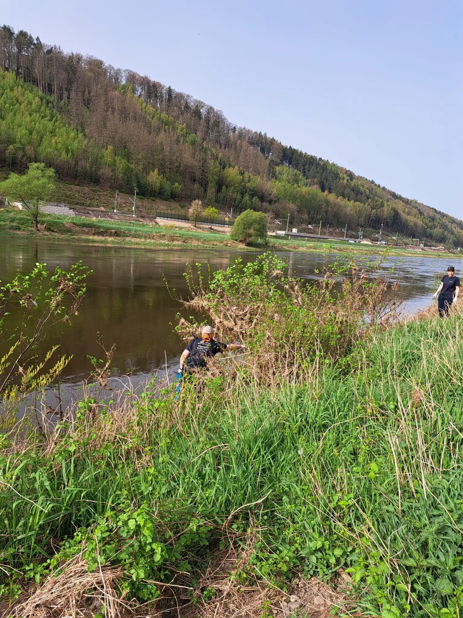 Eine Naturlandschaft mit einem Fluss und einem bewaldeten Hang im Hintergrund, auf der zwei Personen am Ufer zu sehen sind.
