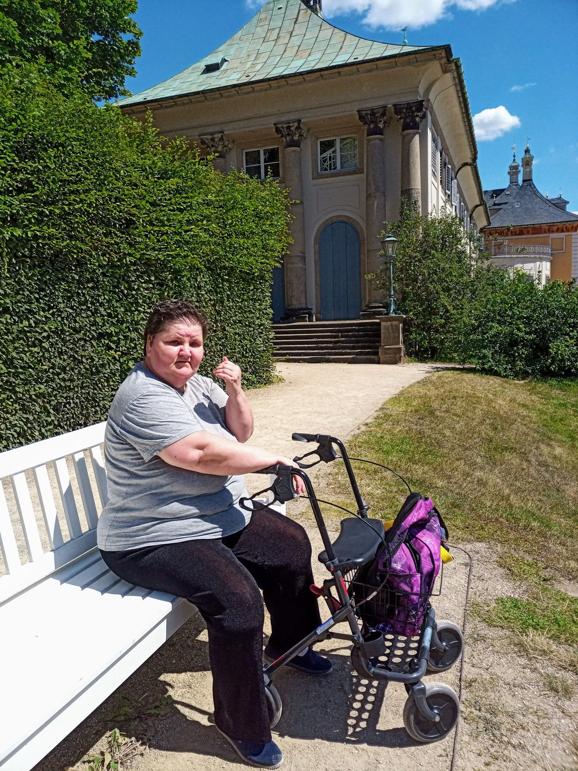 Eine Frau sitzt auf einer weißen Parkbank und hält einen Rollator, im Hintergrund ist ein historisches Gebäude unter blauem Himmel zu sehen.