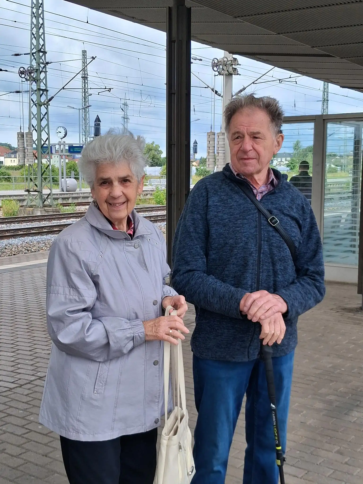 Ein älteres Paar steht lächelnd an einem Bahnsteig. Der Mann hält einen Gehstock.
