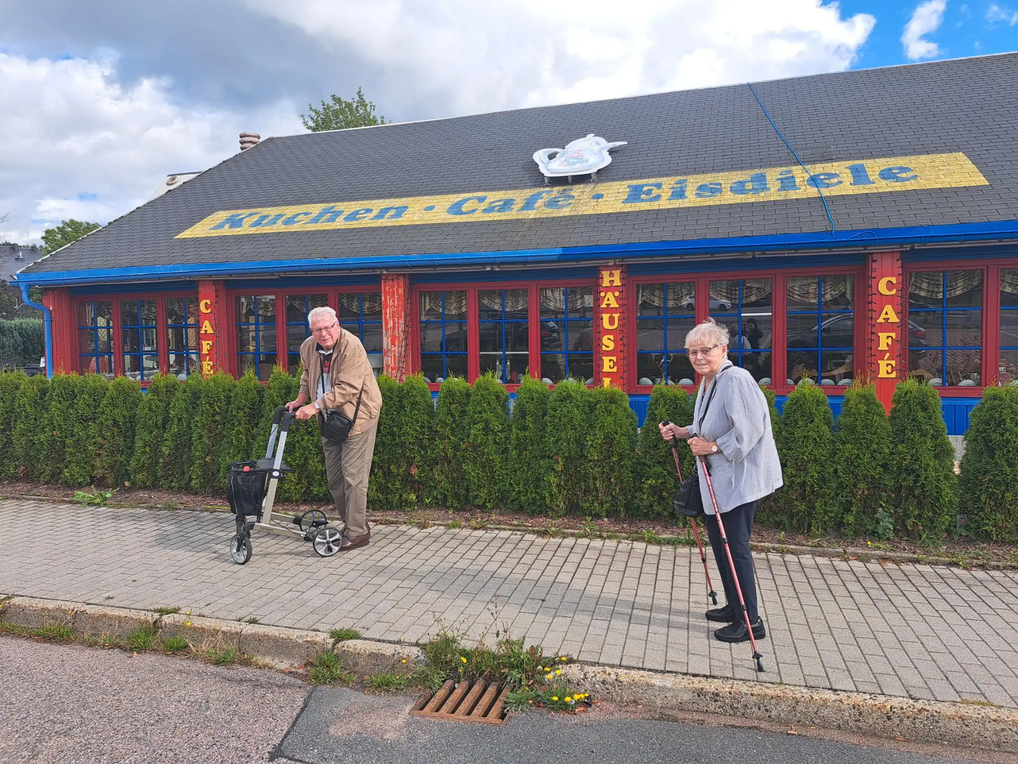Zwei ältere Menschen mit Gehhilfen stehen lächelnd auf einem Gehweg vor einem Cafégebäude.