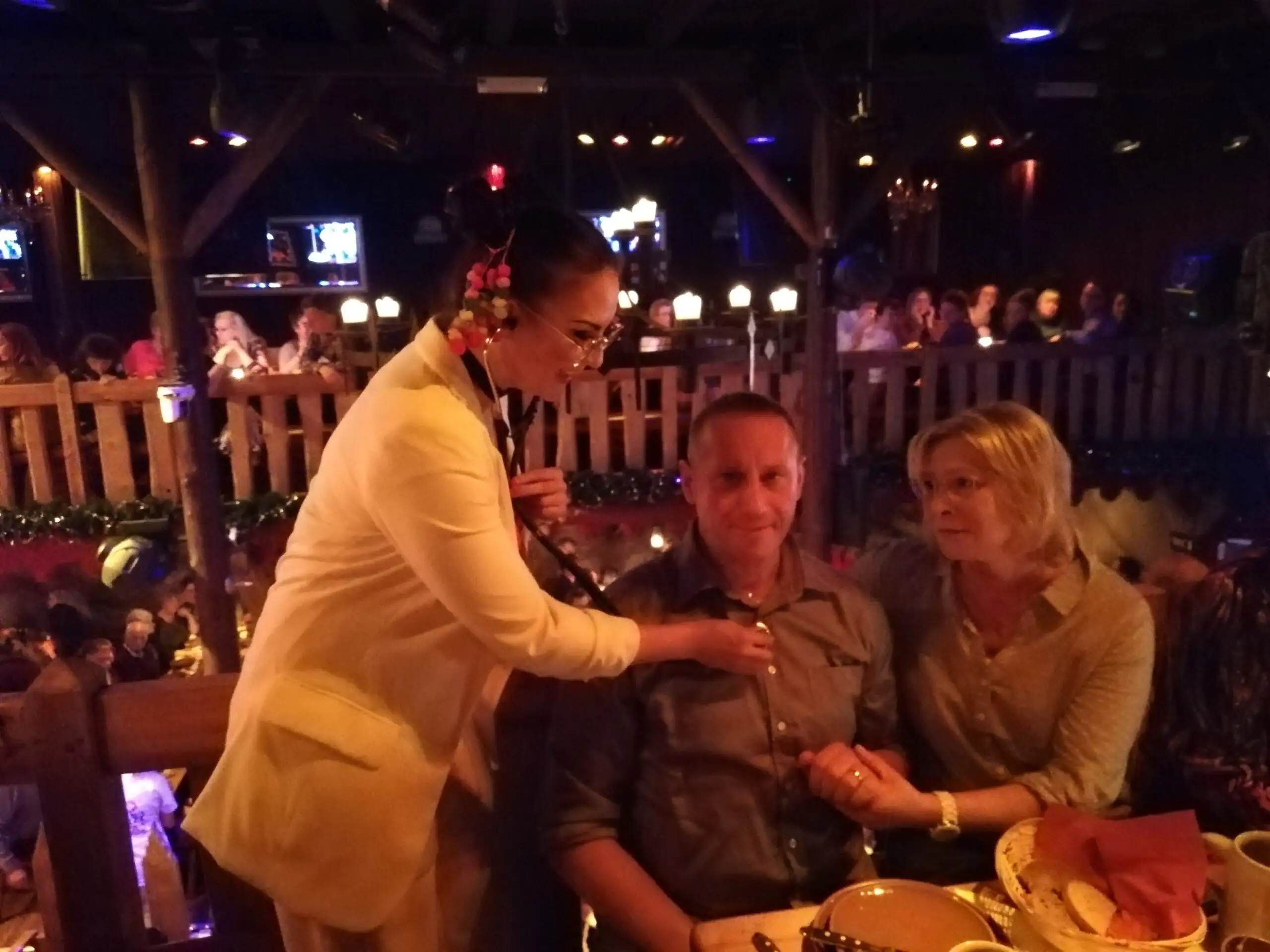 Eine Frau in heller Jacke steht an einem Tisch und interagiert mit einem sitzenden Paar in einem belebten Restaurant mit gedimmtem Licht.