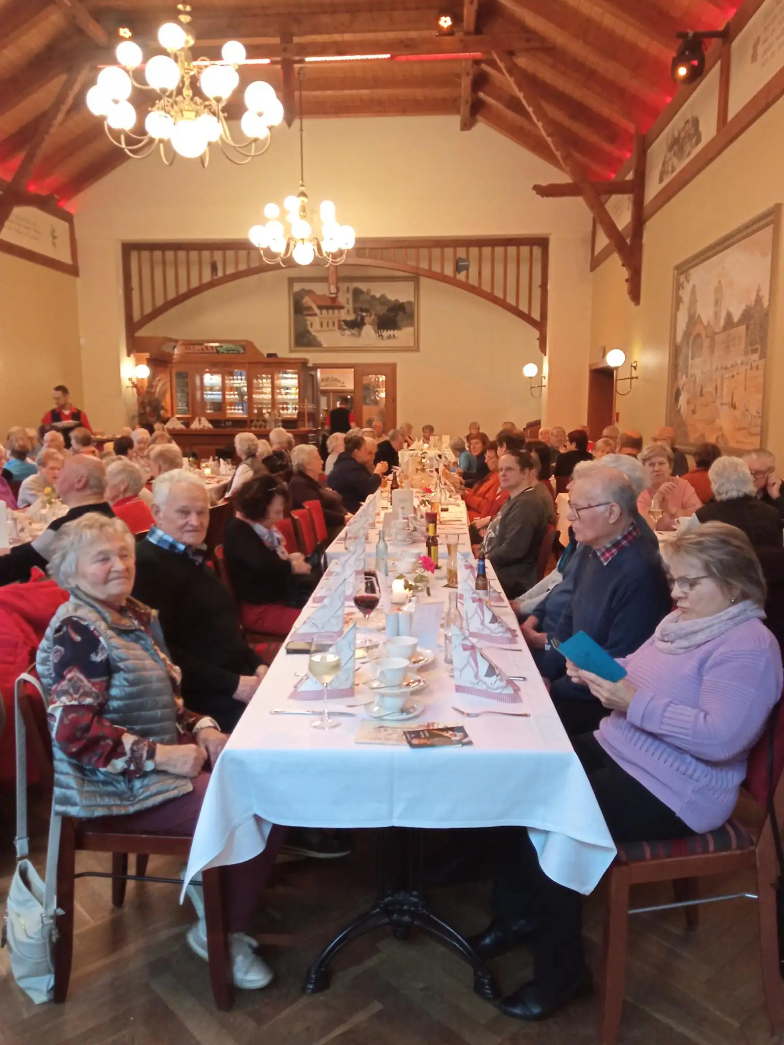 Eine Gruppe älterer Menschen sitzt an einem langen gedeckten Tisch bei einer gemeinsamen Mahlzeit in einem großen Saal.