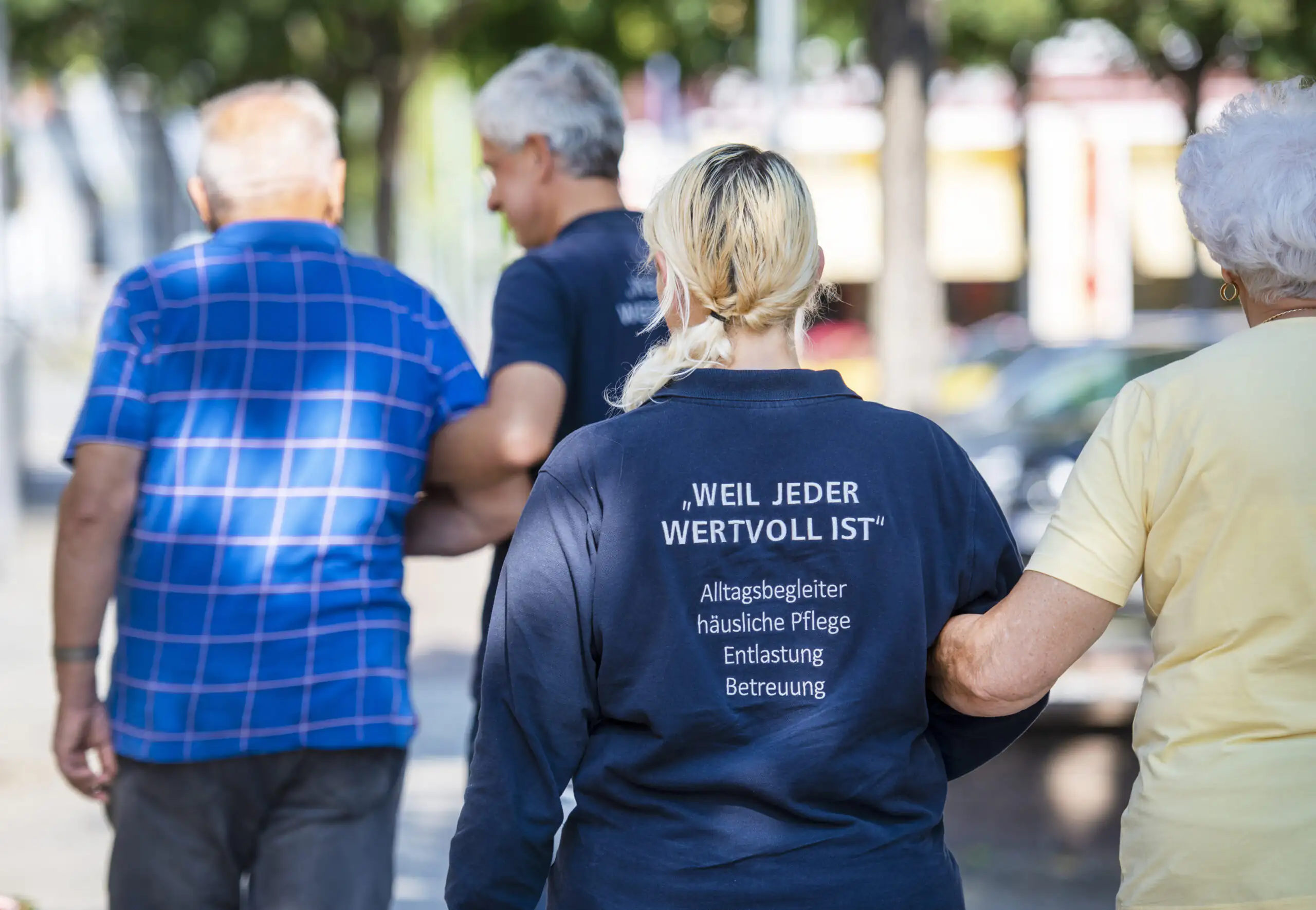 Eine Pflegekraft begleitet zwei ältere Menschen beim Spaziergang im Freien. Die Gruppe bewegt sich gemeinsam weg von der Kamera.