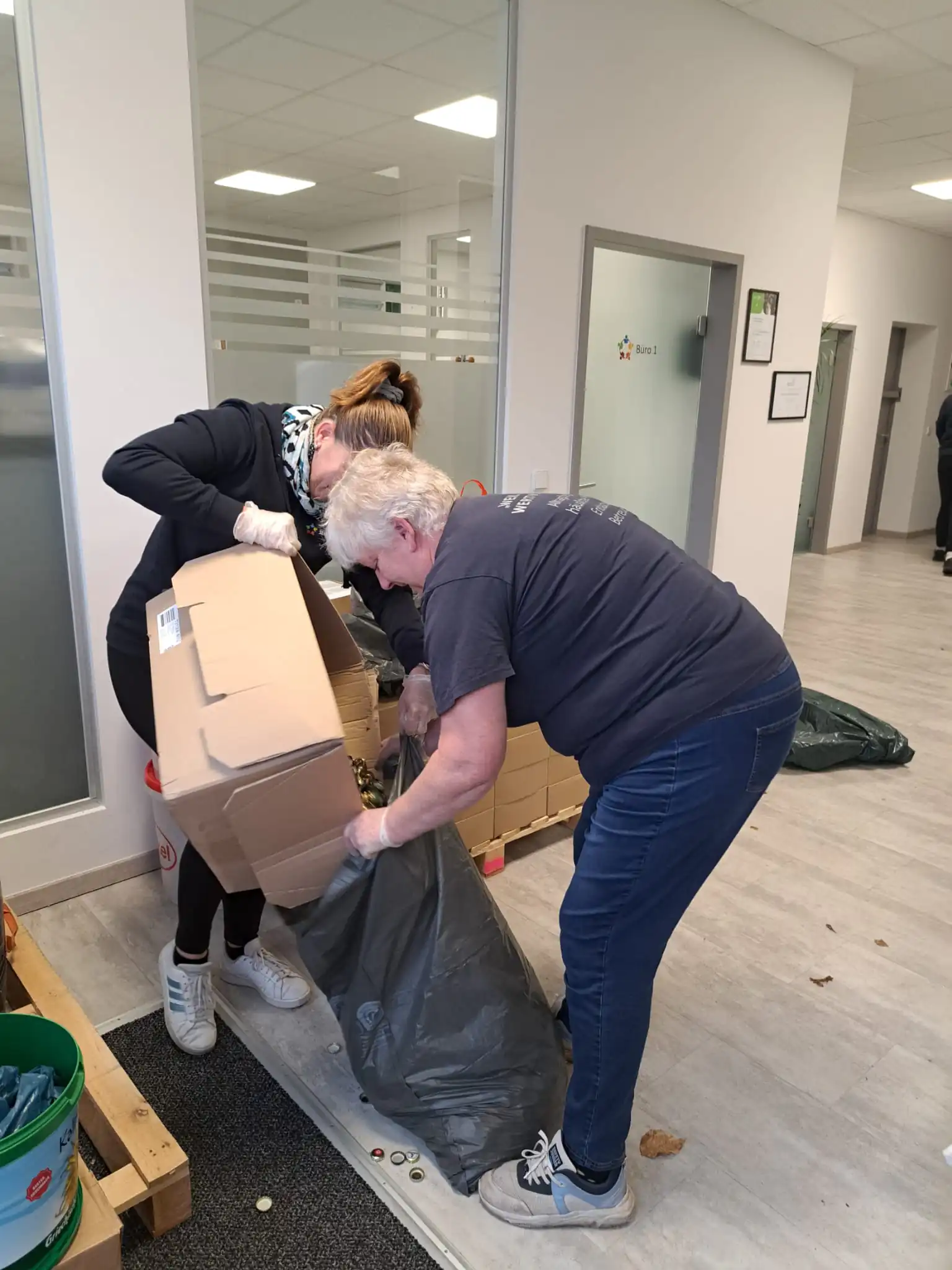 Zwei Frauen arbeiten gemeinsam bei Aufräumarbeiten in einem hellen Büroflur und füllen Kartons in einen Müllbeutel.