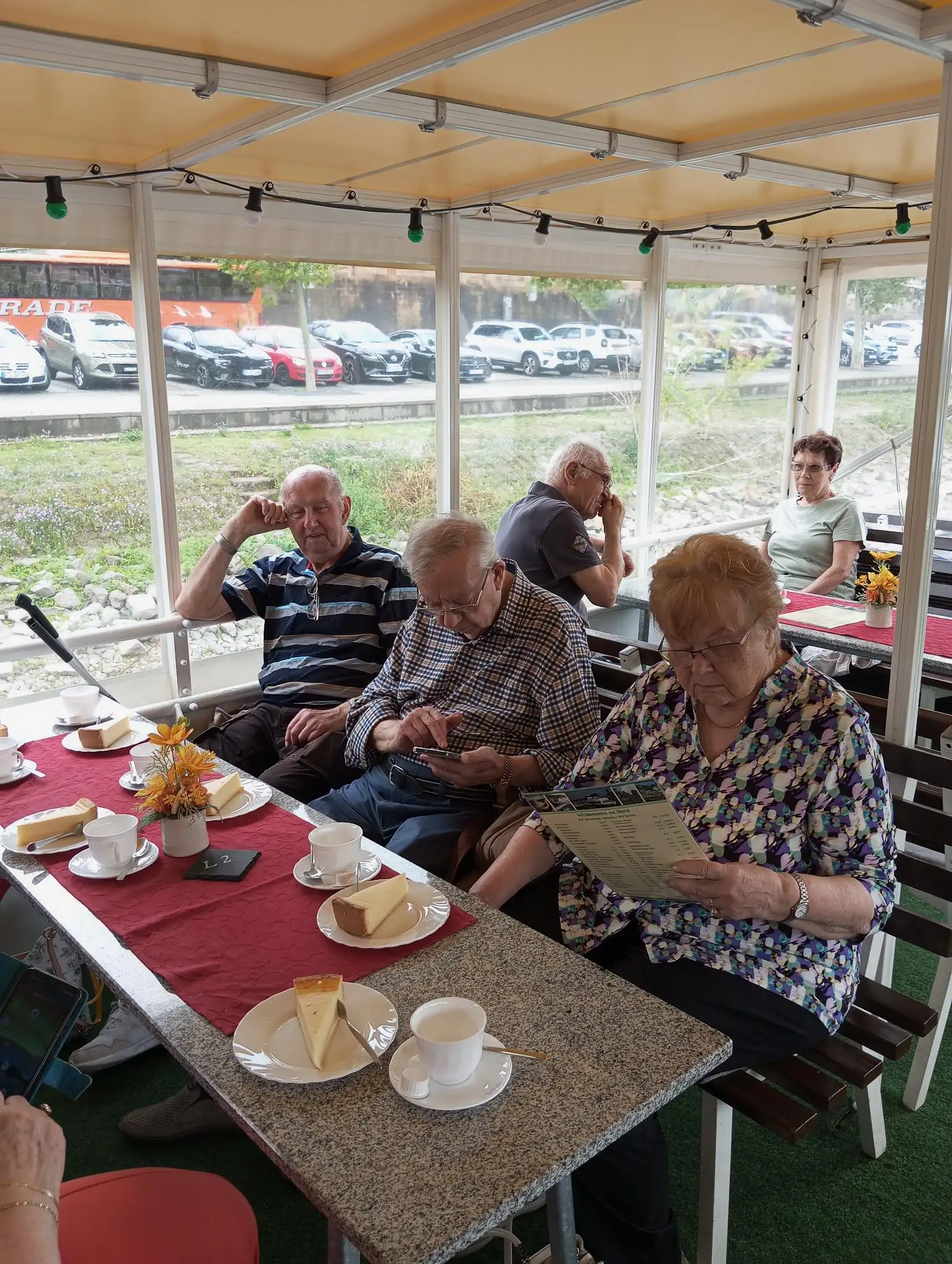 Eine Gruppe älterer Menschen sitzt an einem gedeckten Tisch und genießt Kaffee und Kuchen. Die Szene zeigt eine entspannte Gemeinschaftssituation in einer überdachten Terrasse.
