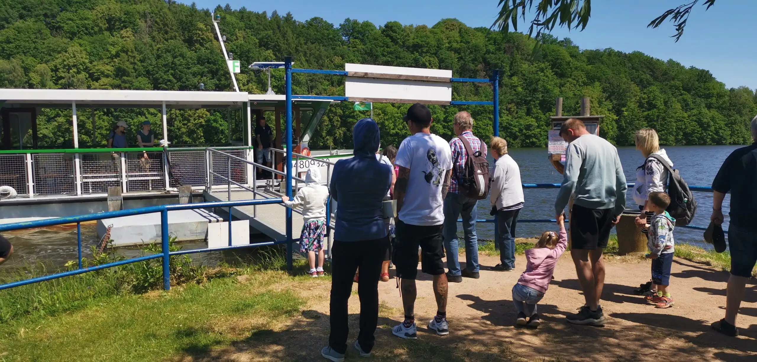 Eine gemischte Gruppe von Menschen wartet an einem sonnigen Ufer auf den Einstieg in ein Fahrgastschiff. Die Szene zeigt eine entspannte Atmosphäre bei einem Ausflug in der Natur.