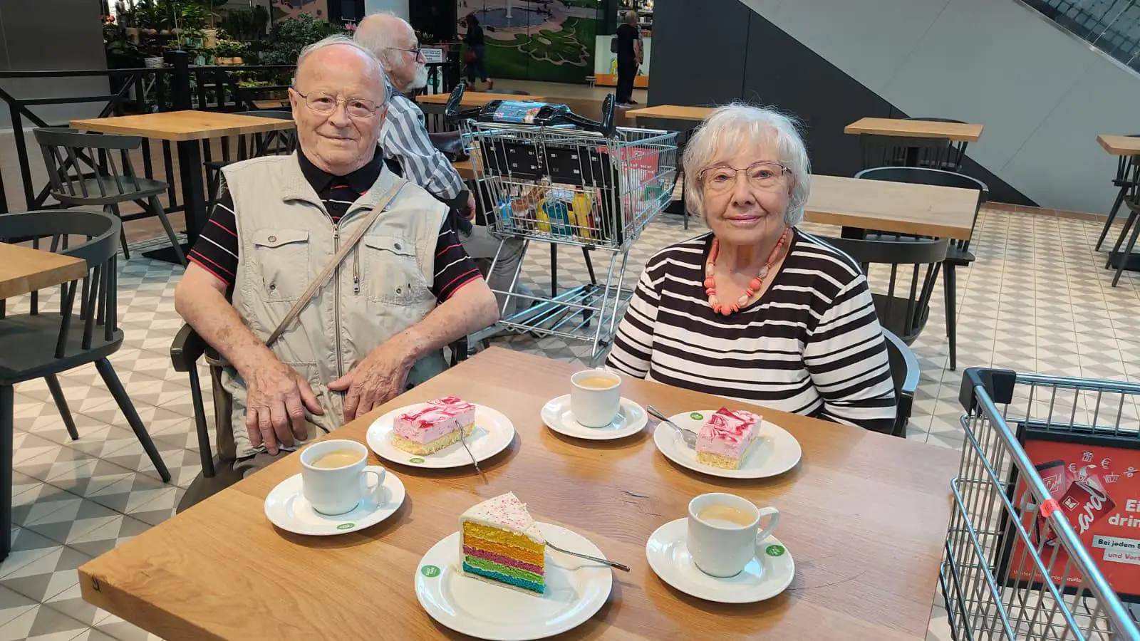 Ein älteres Ehepaar sitzt an einem Tisch und genießt Kaffee und Kuchen in einem öffentlichen Café-Bereich.