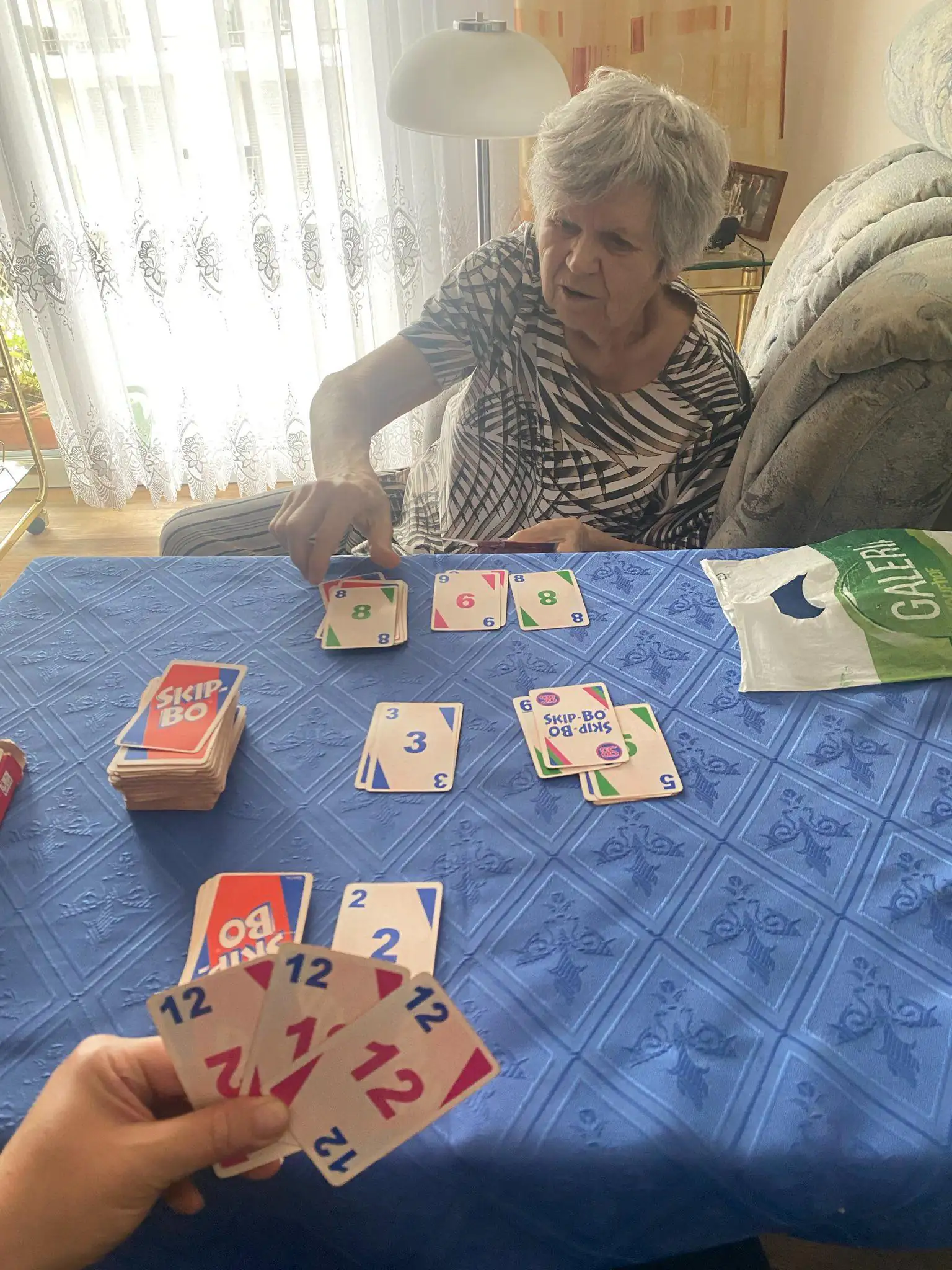 Eine ältere Dame spielt ein Kartenspiel am Tisch in einem hellen Raum. Im Vordergrund ist die Hand einer zweiten Person zu sehen.