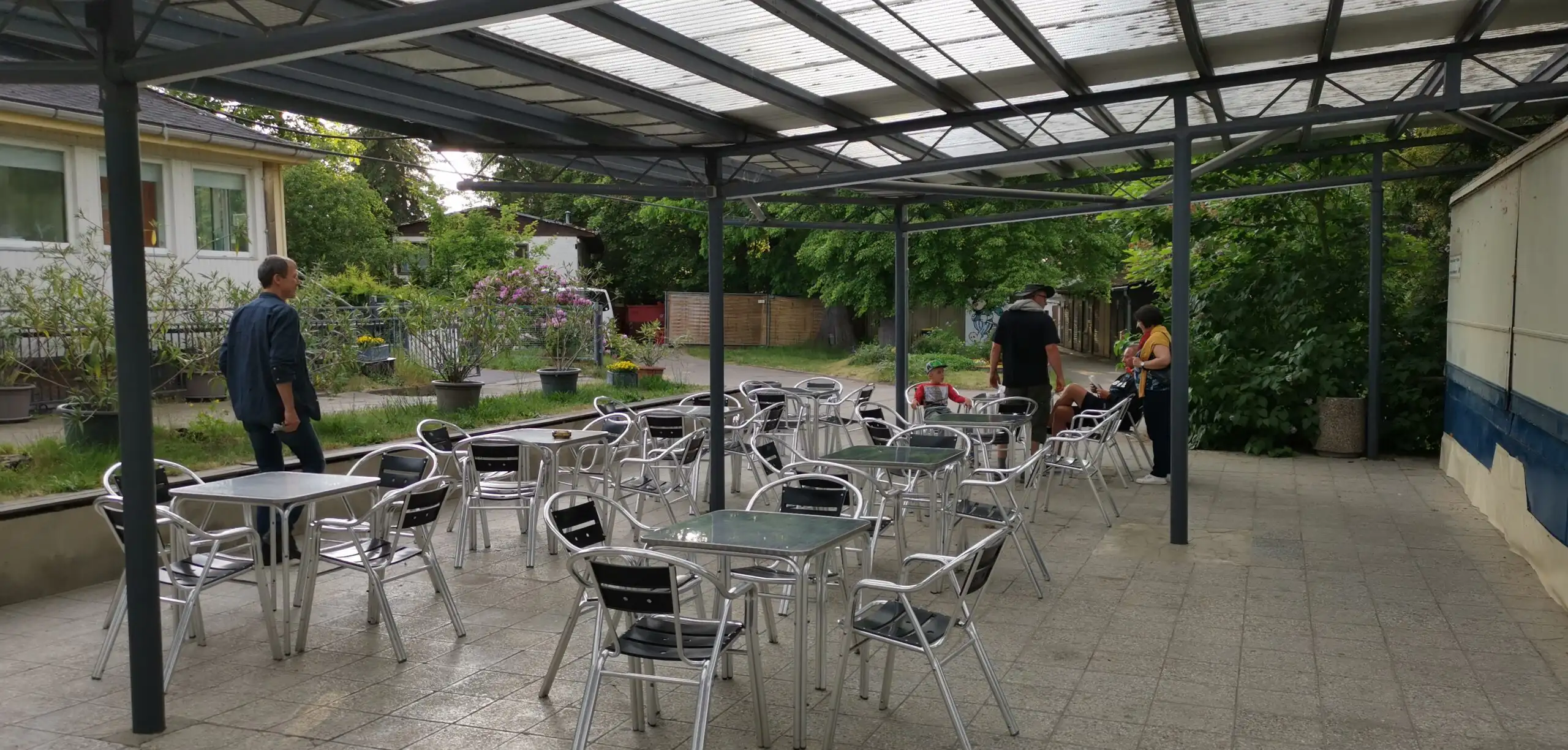Ein überdachter Außenbereich mit vielen Tischen und Stühlen, der als Terrasse oder Gartenbereich dient. Im Hintergrund sind Personen und Grünanlagen zu sehen.