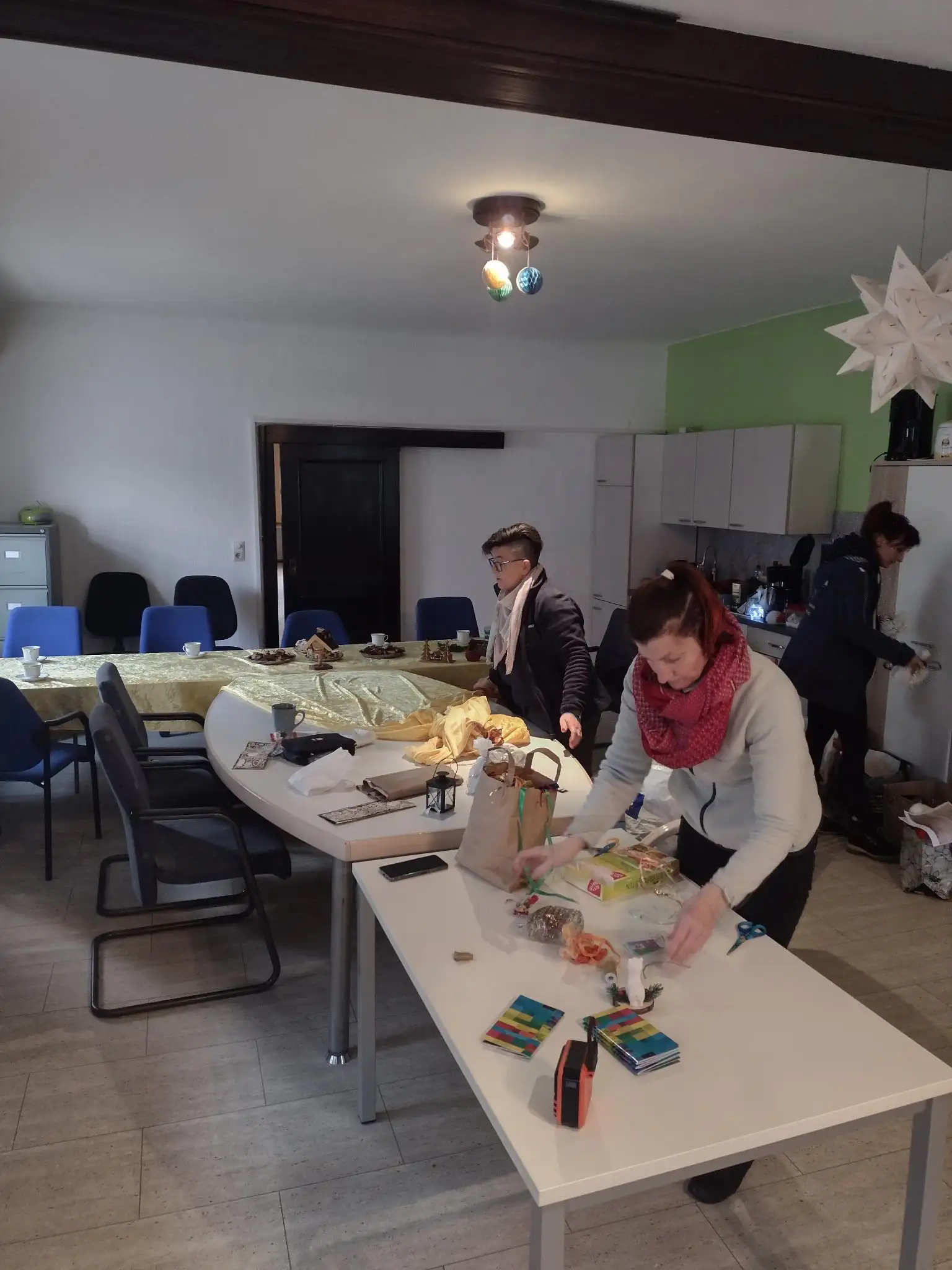 Personen richten Tische in einem Gemeinschaftsraum für eine Veranstaltung ein. Im Hintergrund ist eine Küchenzeile und eine Weihnachtsdekoration zu sehen.