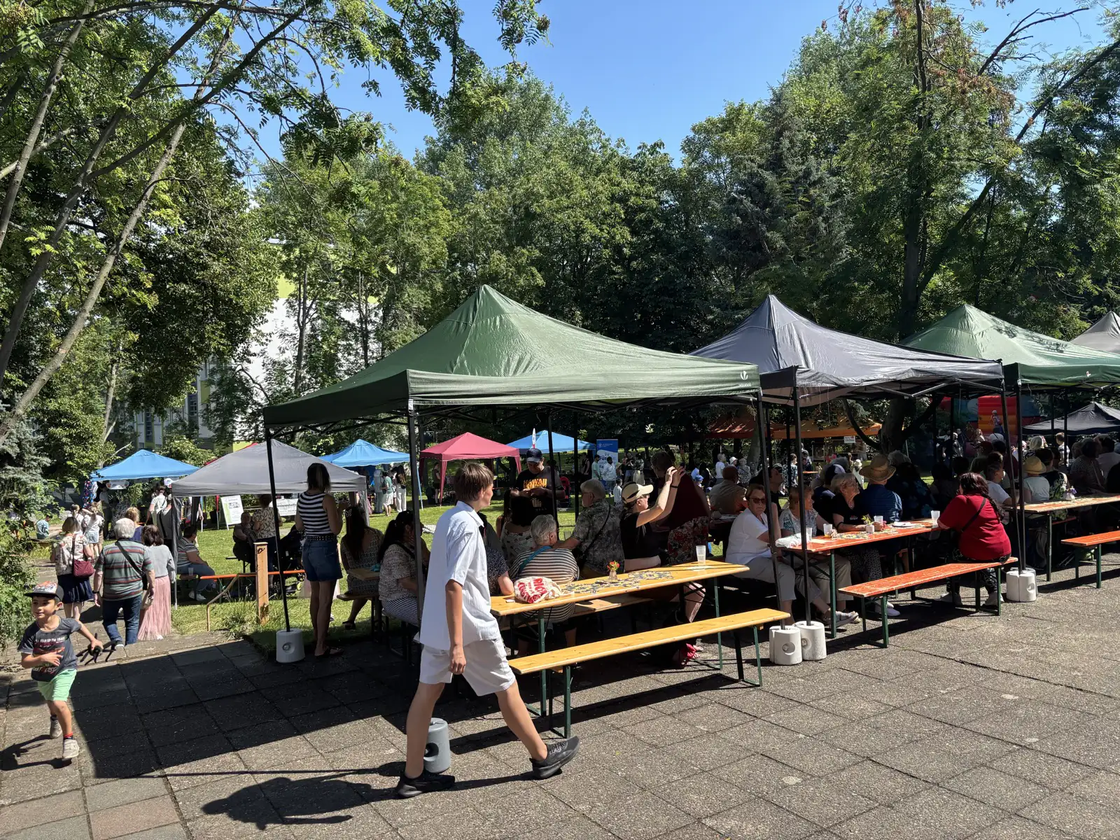 Sonniges Freiluftfest mit Zelten und Besuchern an Tischen im Park. Die Szene zeigt eine gesellige Atmosphäre im Außenbereich mit vielen Menschen.