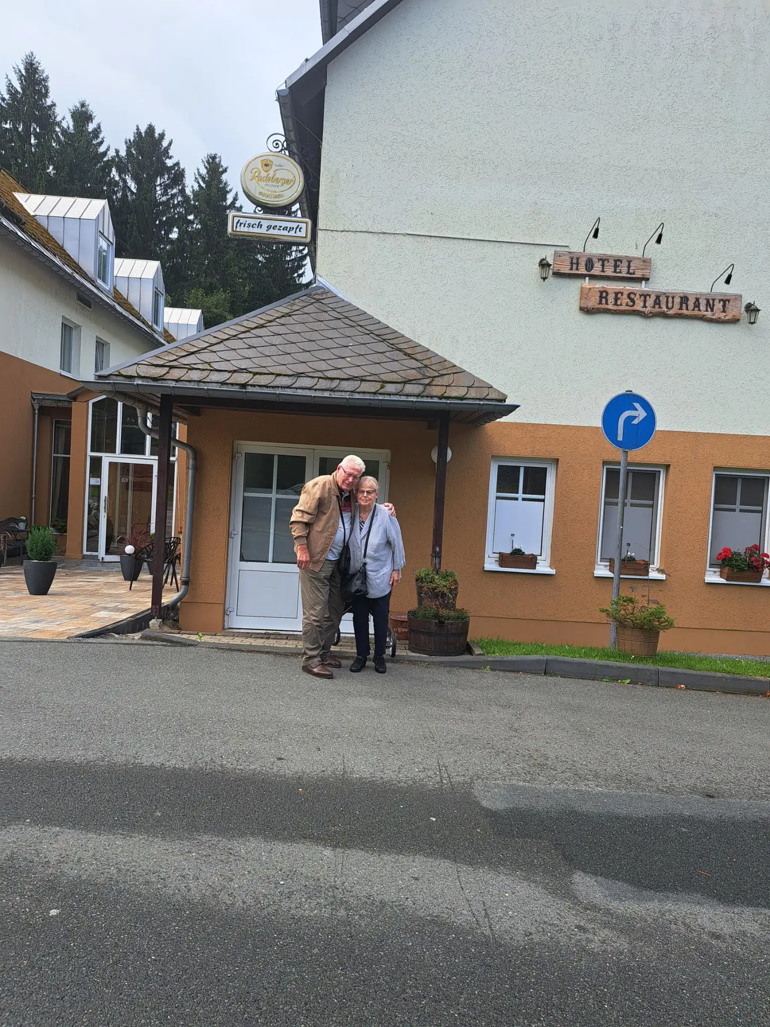 Ein älteres Paar steht lächelnd vor dem Eingang eines Gebäudes mit Hotel- und Restaurantbeschilderung. Die Szene zeigt Senioren in einer Freizeitumgebung im Freien.