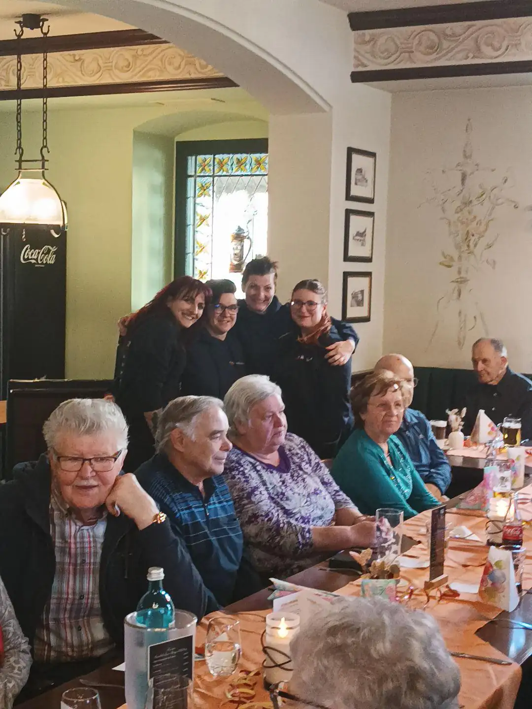Gruppenfoto von Senioren und Pflegepersonal an einem gedeckten Tisch in einem Innenraum. Die Personen wirken entspannt und unterhalten sich bei Getränken und Speisen.